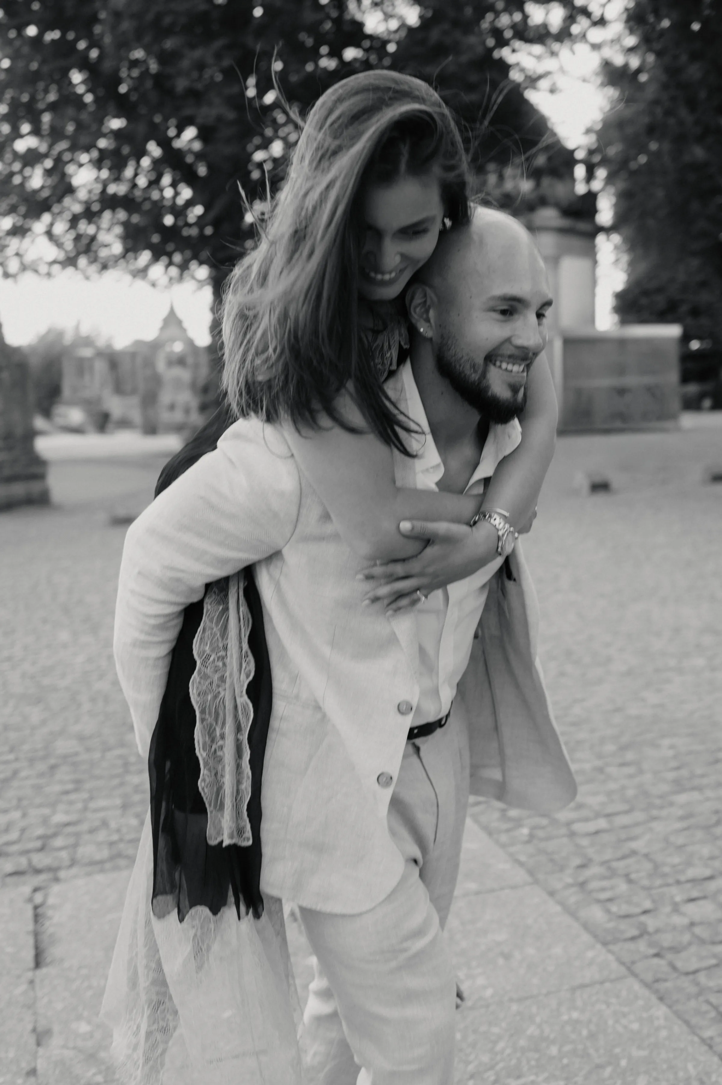 Black-and-white outdoor portrait of a couple sharing a piggyback ride on a cobblestone path.