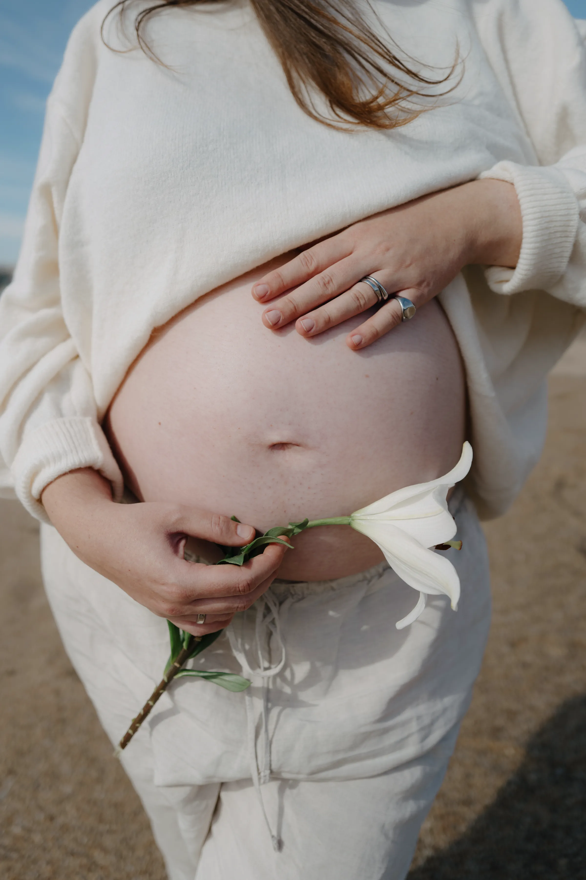 Close portrait of a pregnant belly with hands holding a white flower in soft outdoor light.