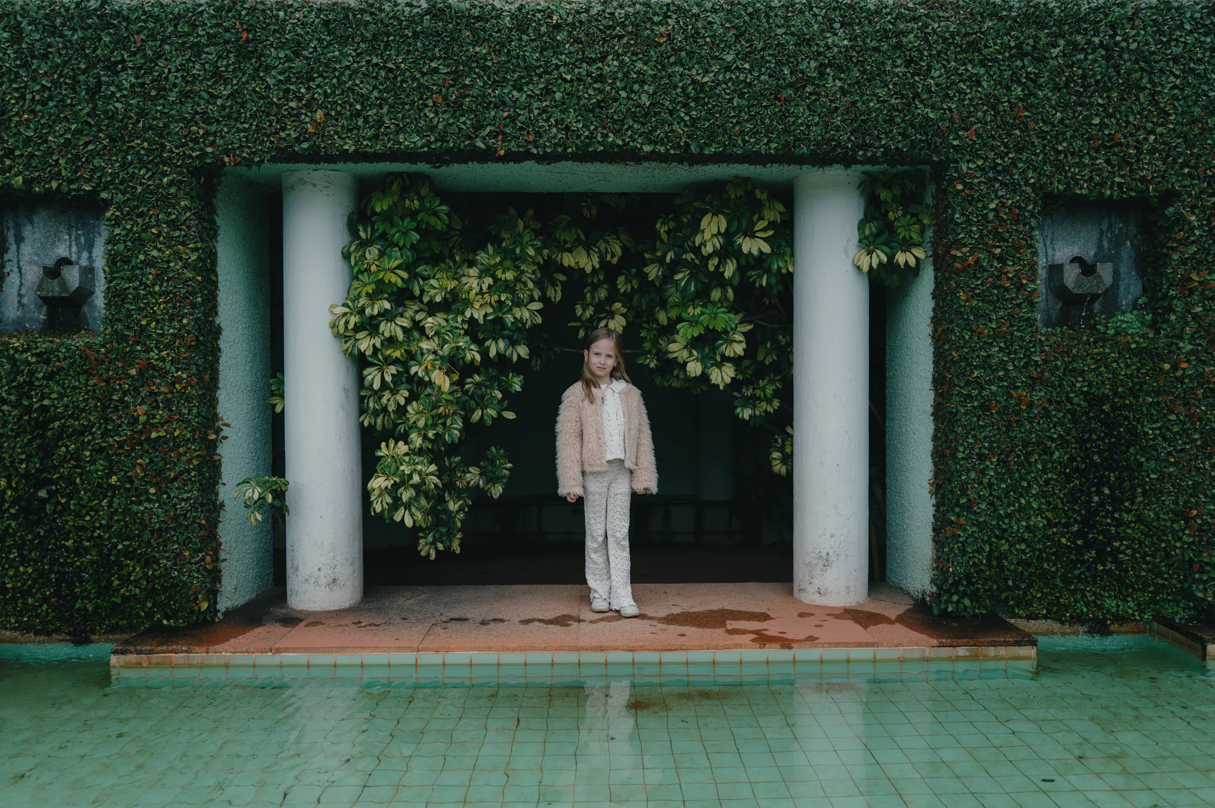 Child portrait by a tiled pool, framed by white columns and dense garden greenery in soft light.