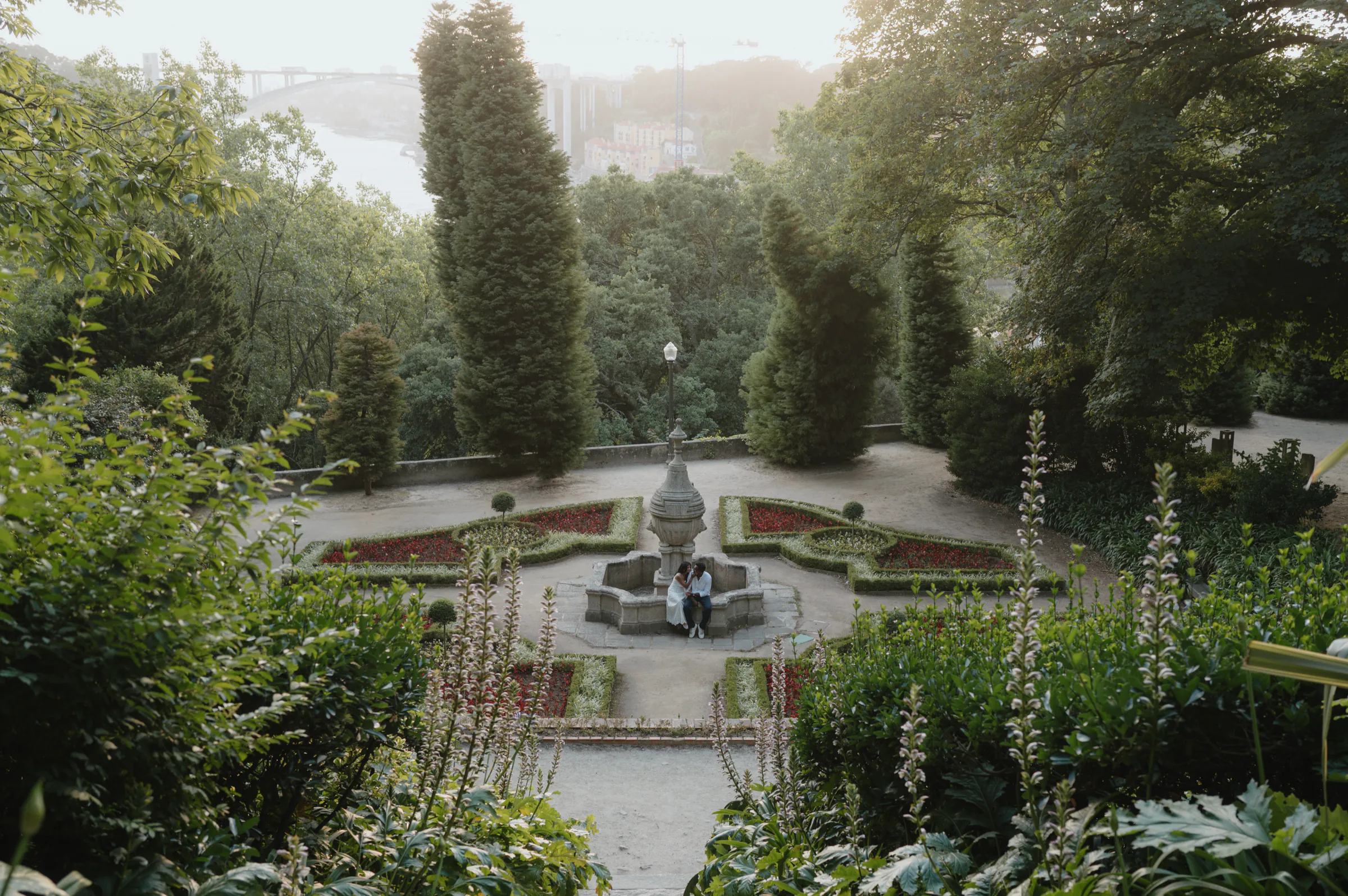 Couple in a formal garden near a fountain, framed by tall trees and soft light.
