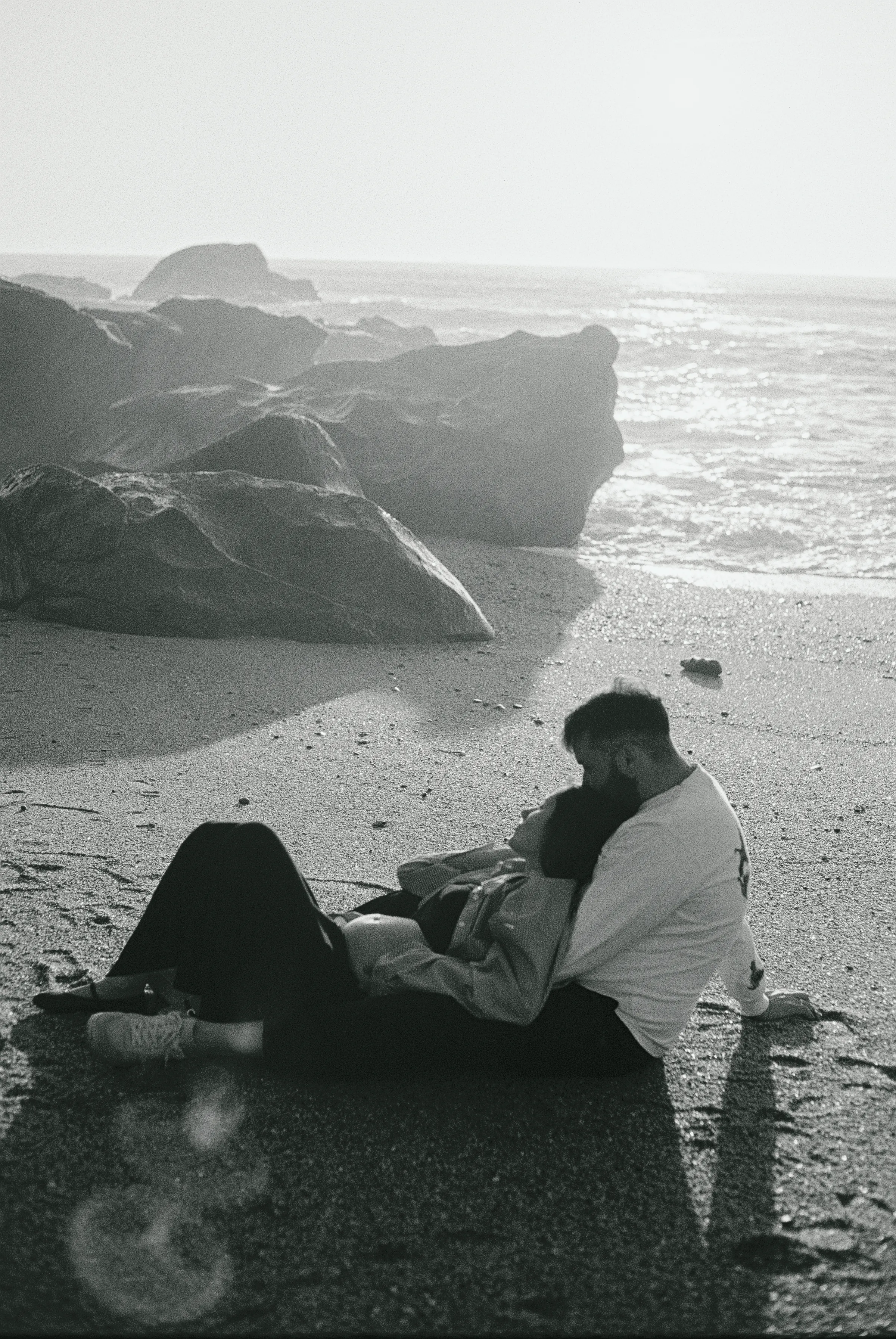 Couple reclining on a rocky seaside beach in soft light, photographed in black and white.