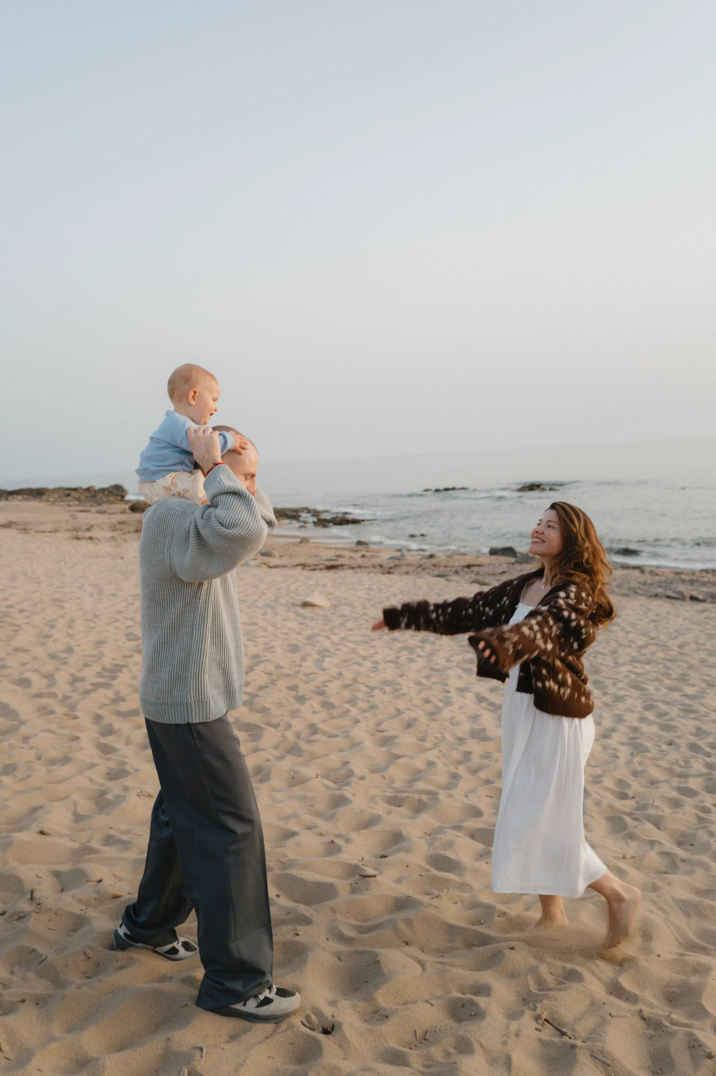 Family seaside session with an adult lifting a baby as another adult reaches out on the sand