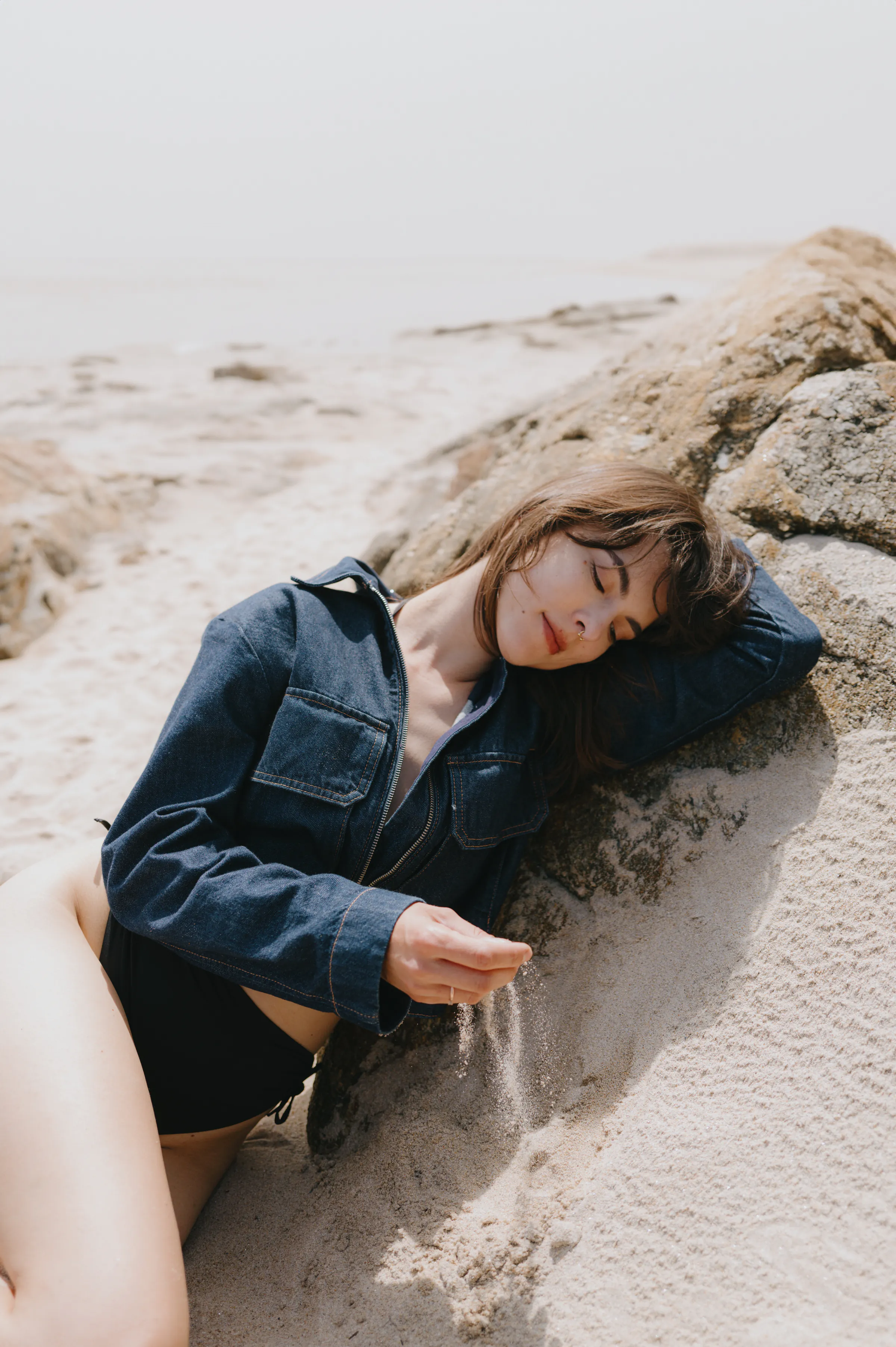 Seaside portrait of a person reclining on sand beside rocks in soft light.
