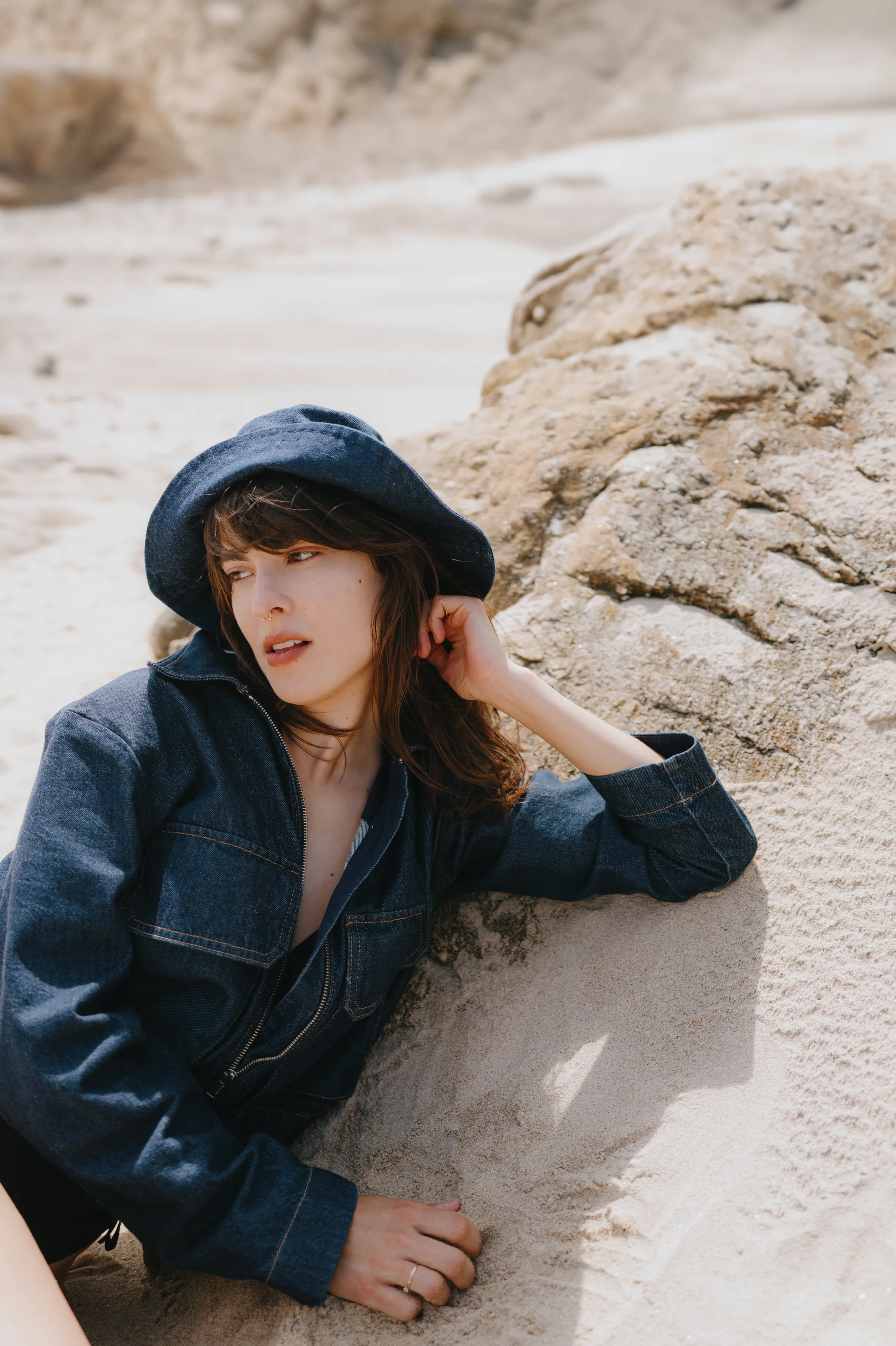 Outdoor portrait of a woman in a dark hat and jacket leaning against pale sandy rock in soft light.