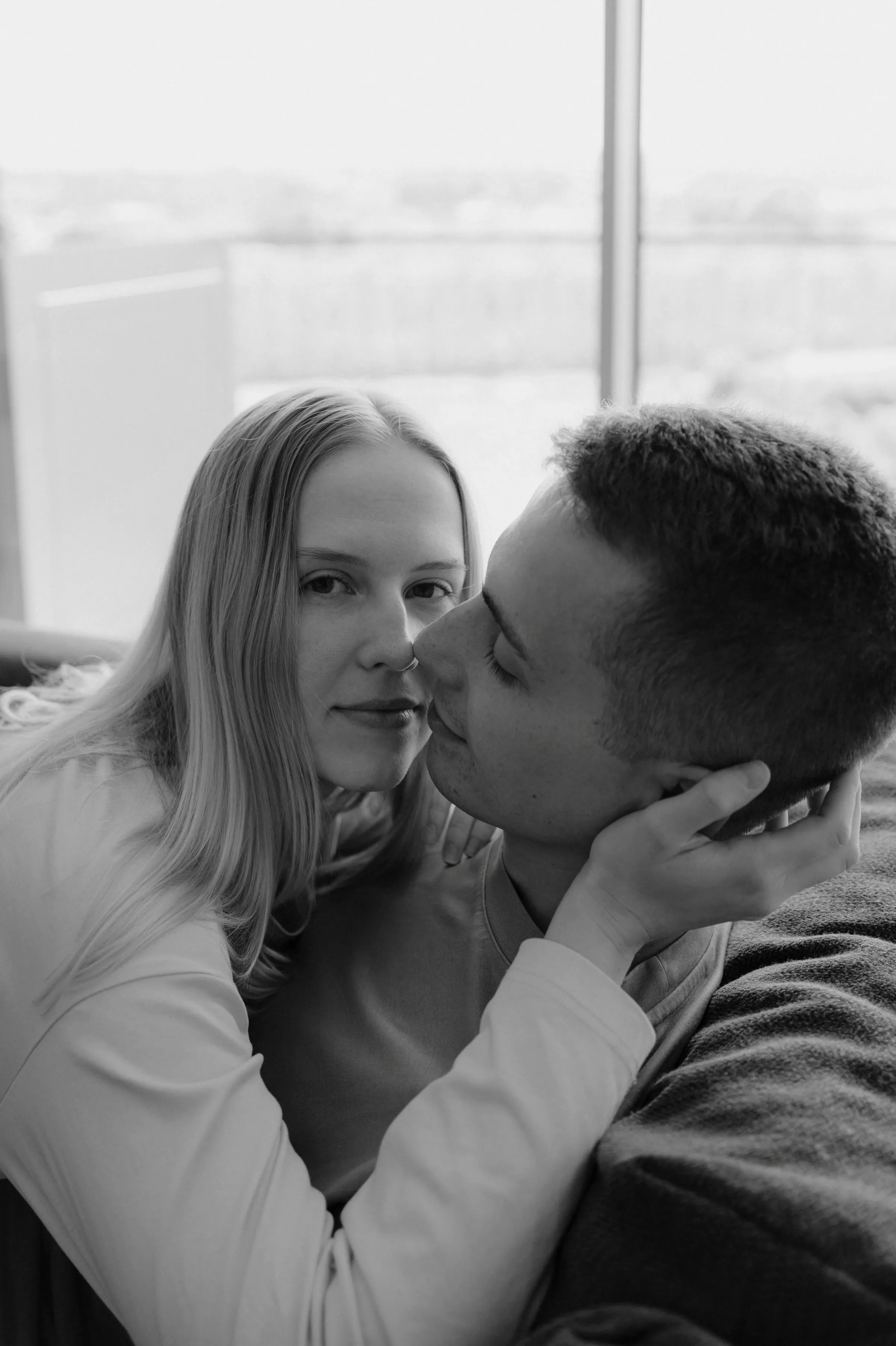 Black-and-white close portrait of a couple seated closely by a window in soft indoor light.