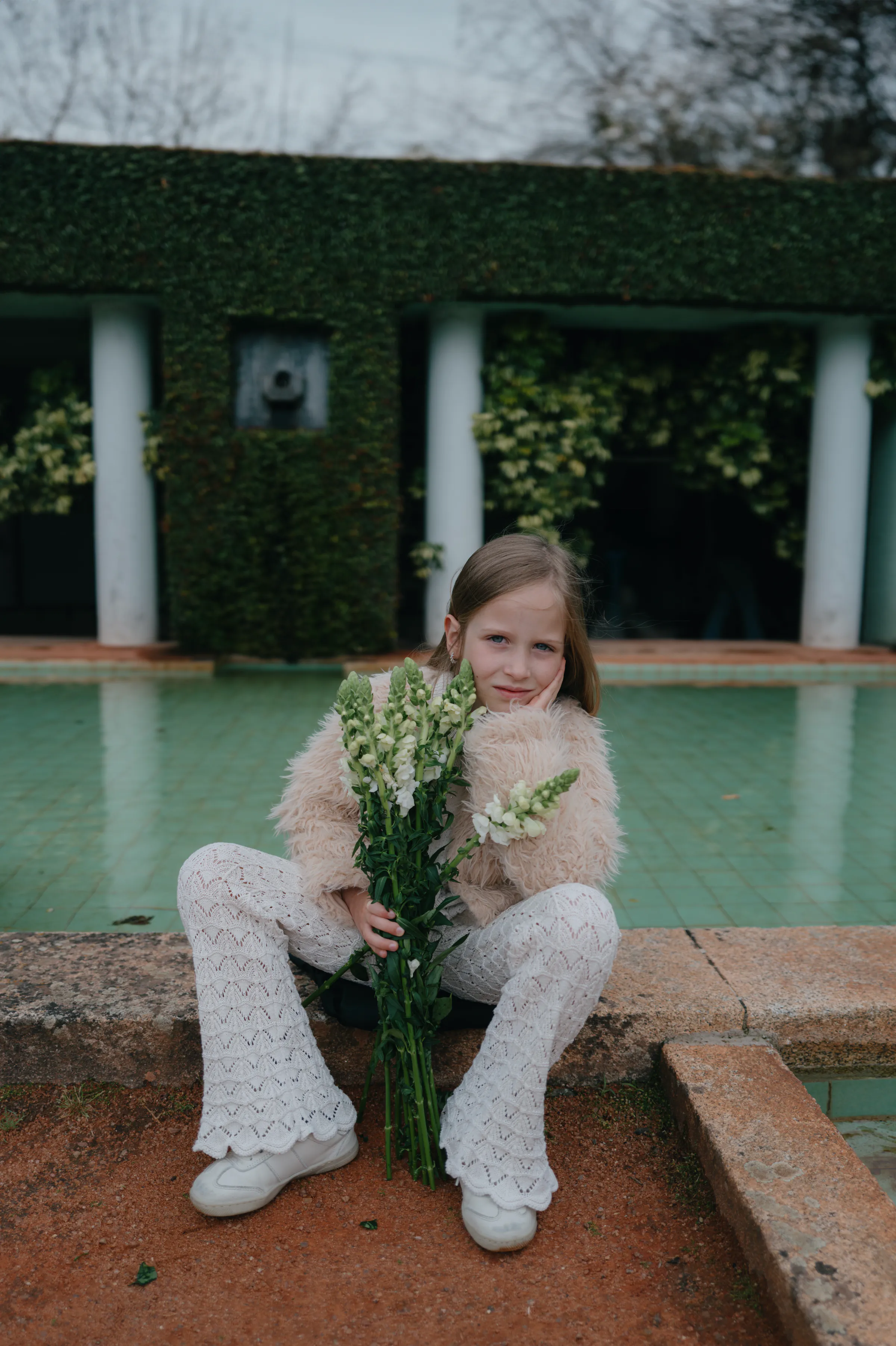 Child portrait by a garden pool, seated with a bouquet in soft outdoor light.