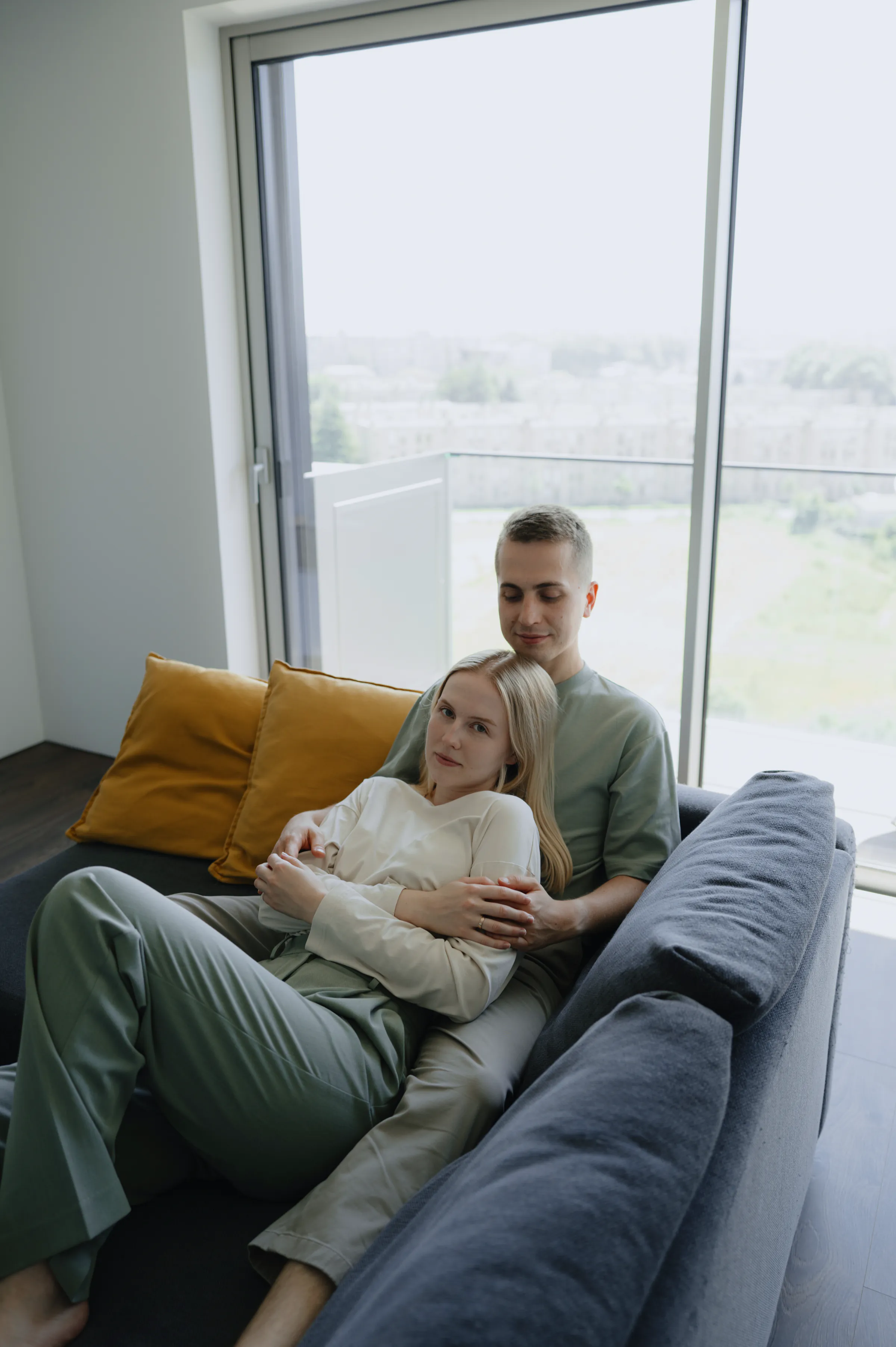 Couple relaxing on a sofa during a soft light home session beside a large window.