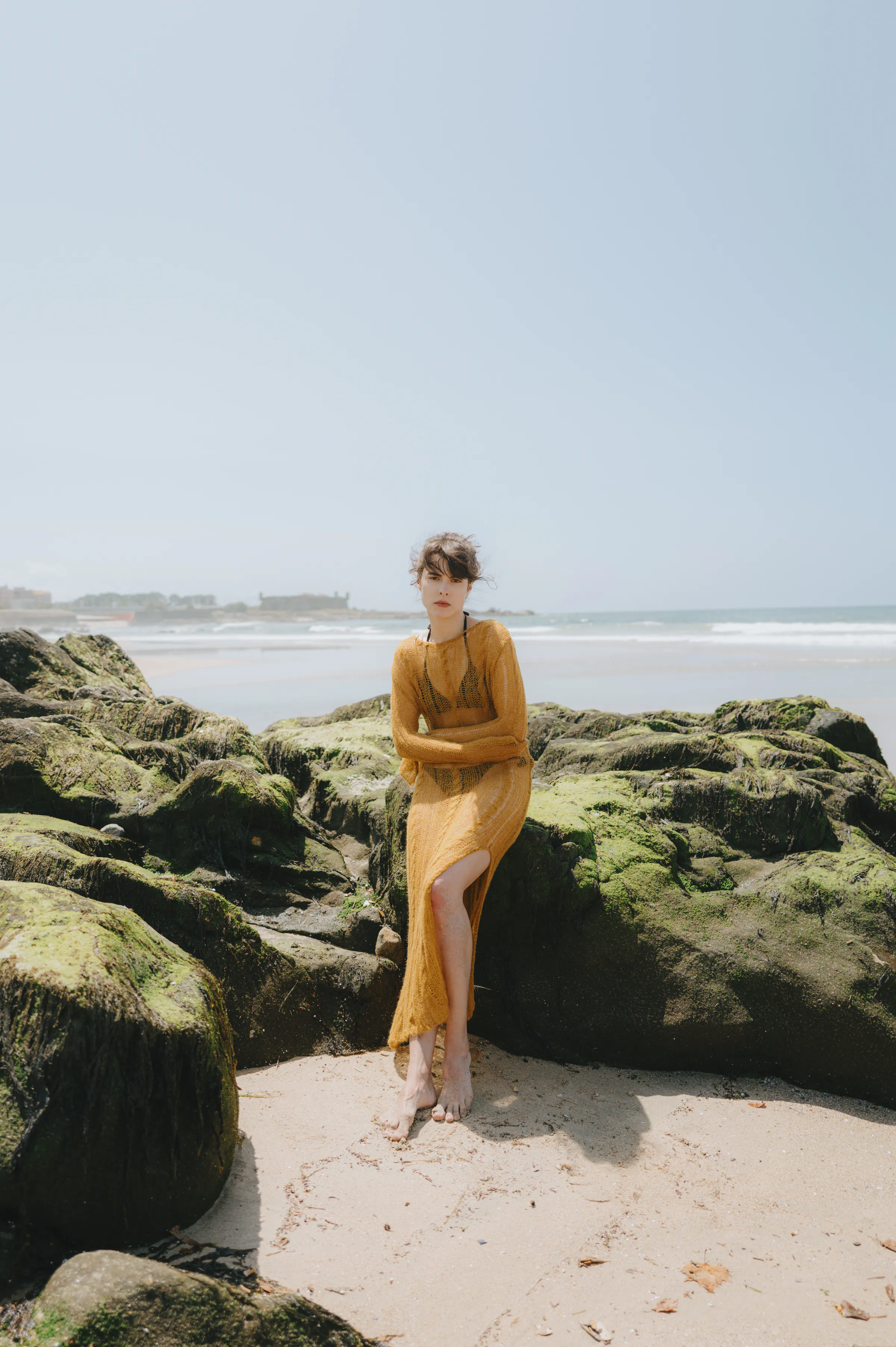 Seaside portrait of an adult seated on mossy rocks in soft light, with waves behind.