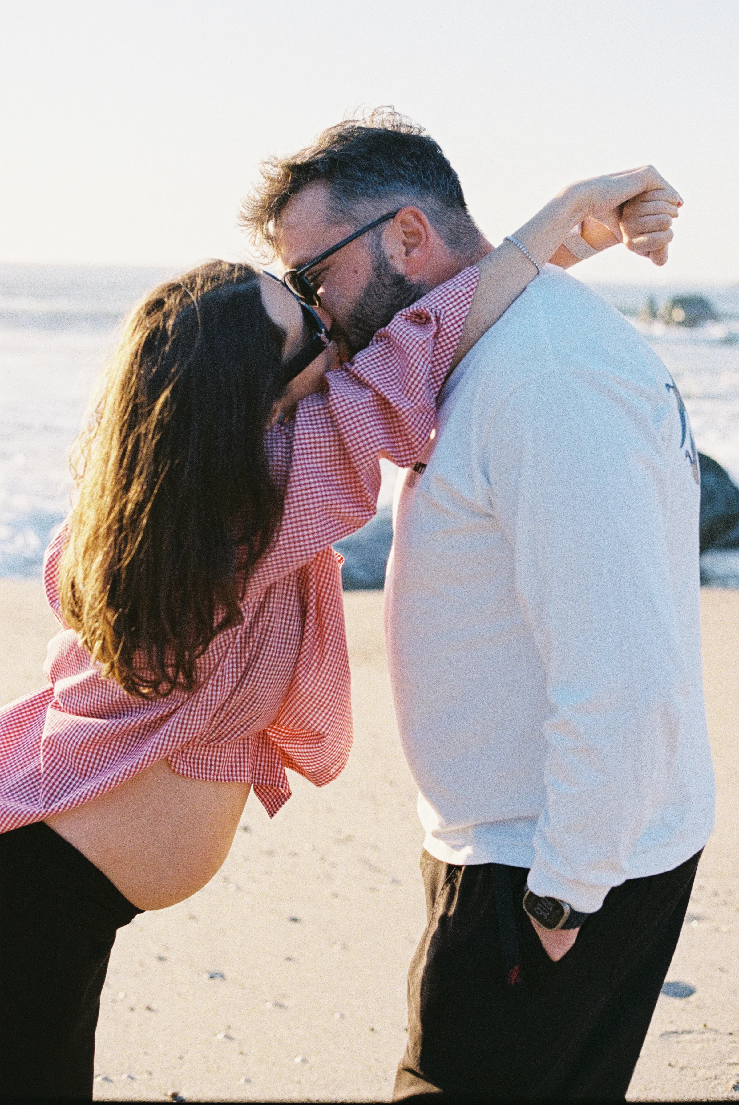 Couple kissing during a seaside outdoor session in soft light, standing close on the sand.