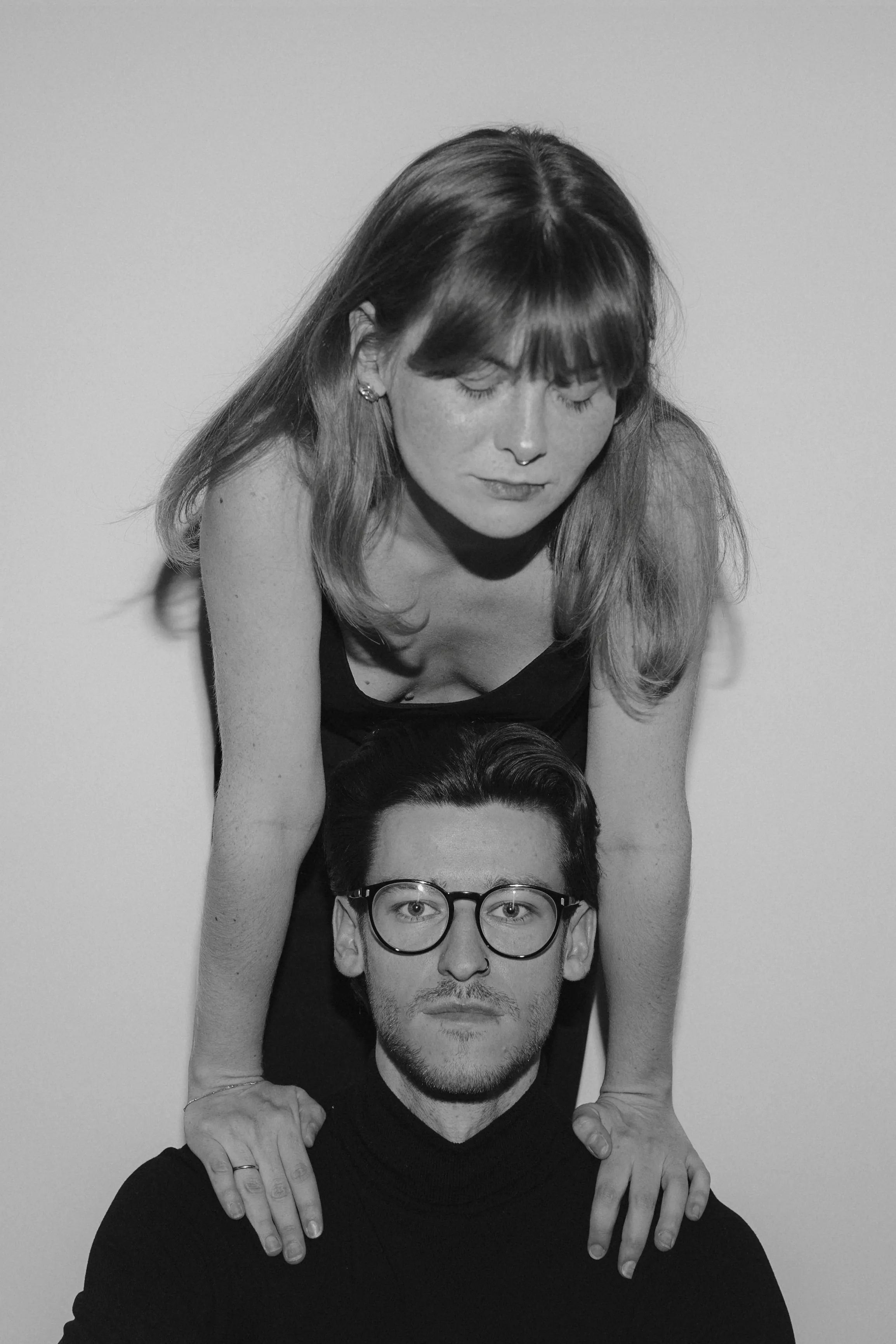 Black-and-white close portrait of a couple against a plain backdrop, one leaning over the other's shoulders.