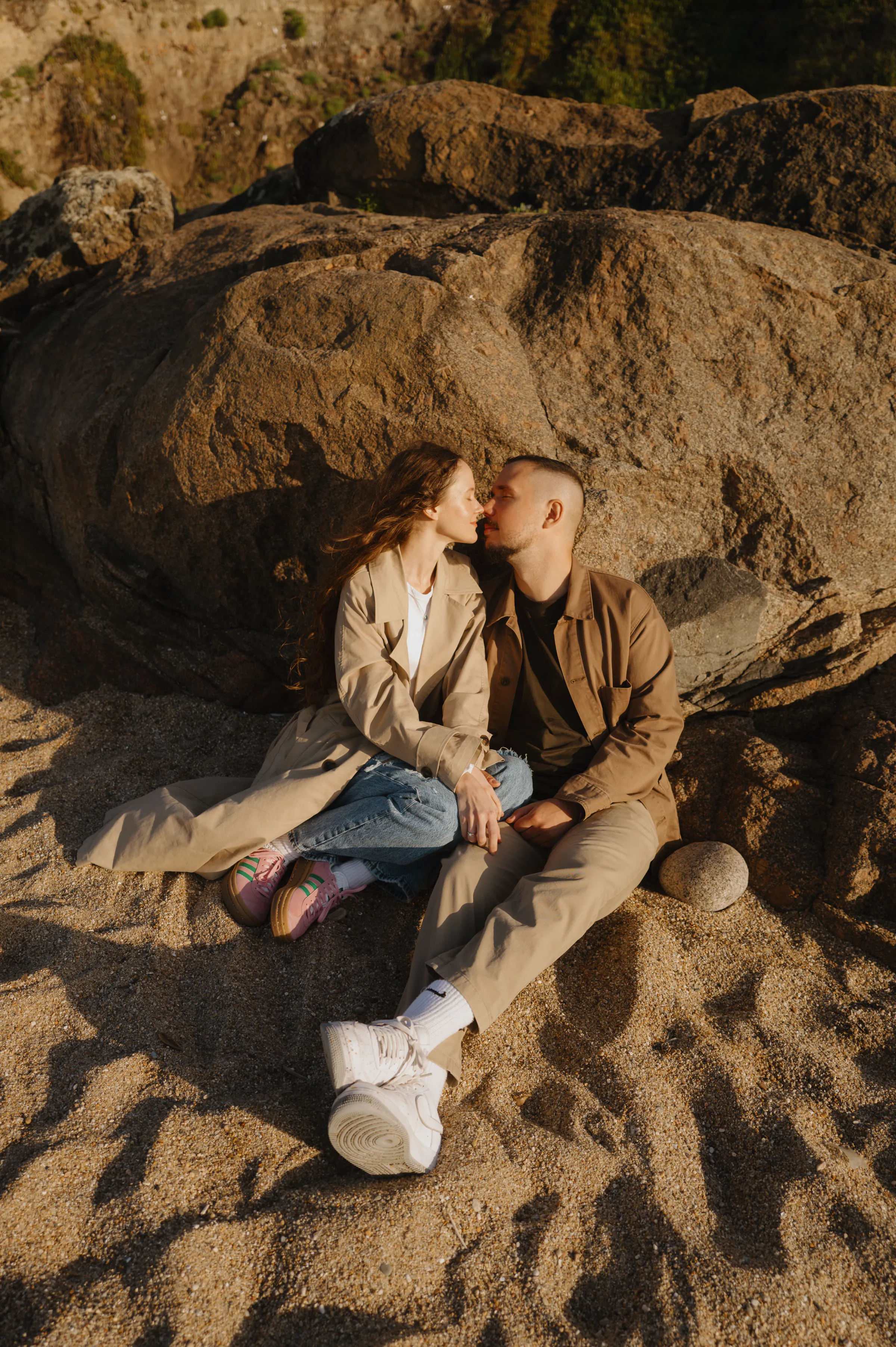 Couple seated on sand beside large rocks, kissing during a seaside outdoor session in warm light.