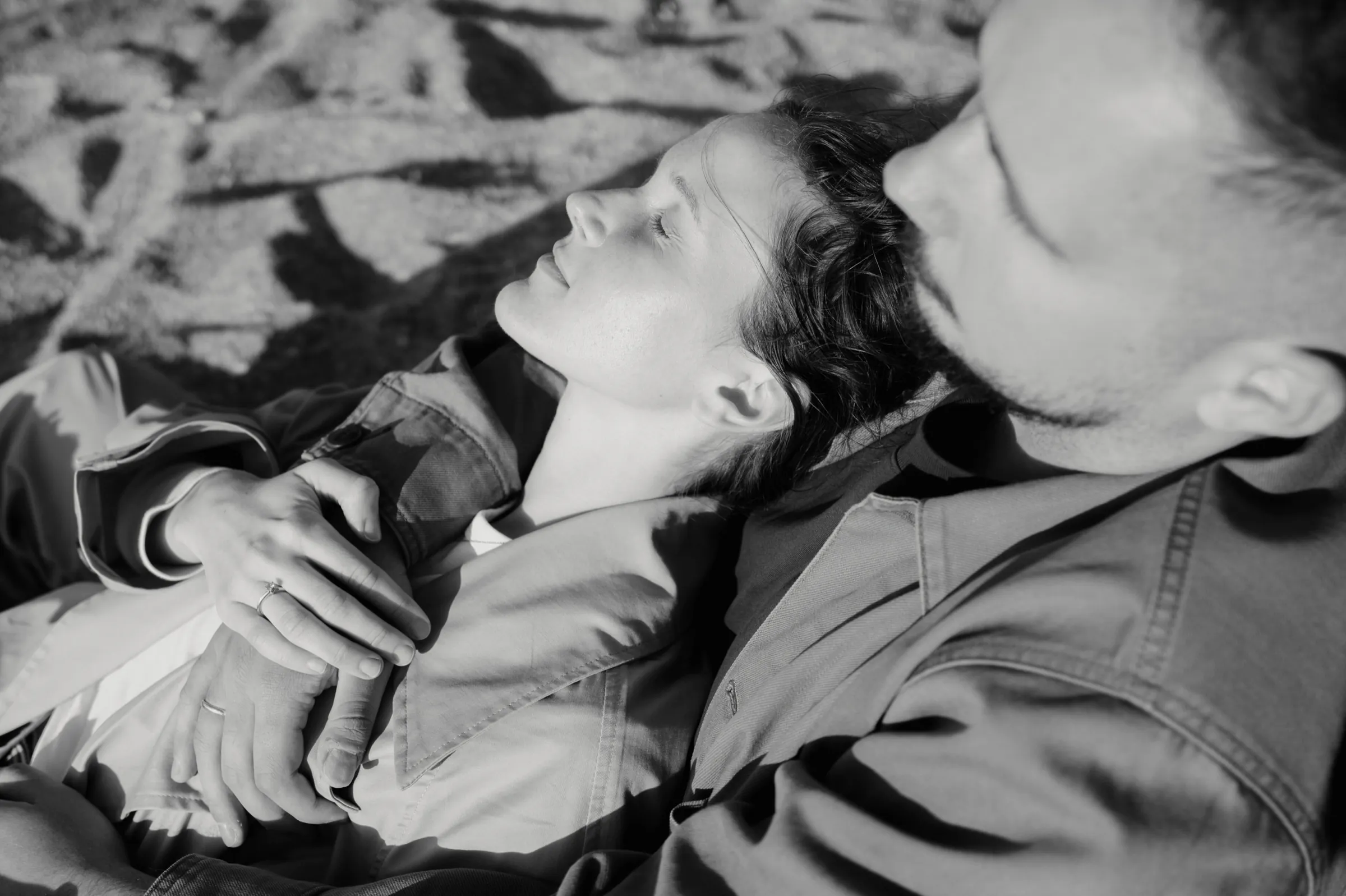 Black-and-white close portrait of a couple lying on sand in soft light, hands intertwined.