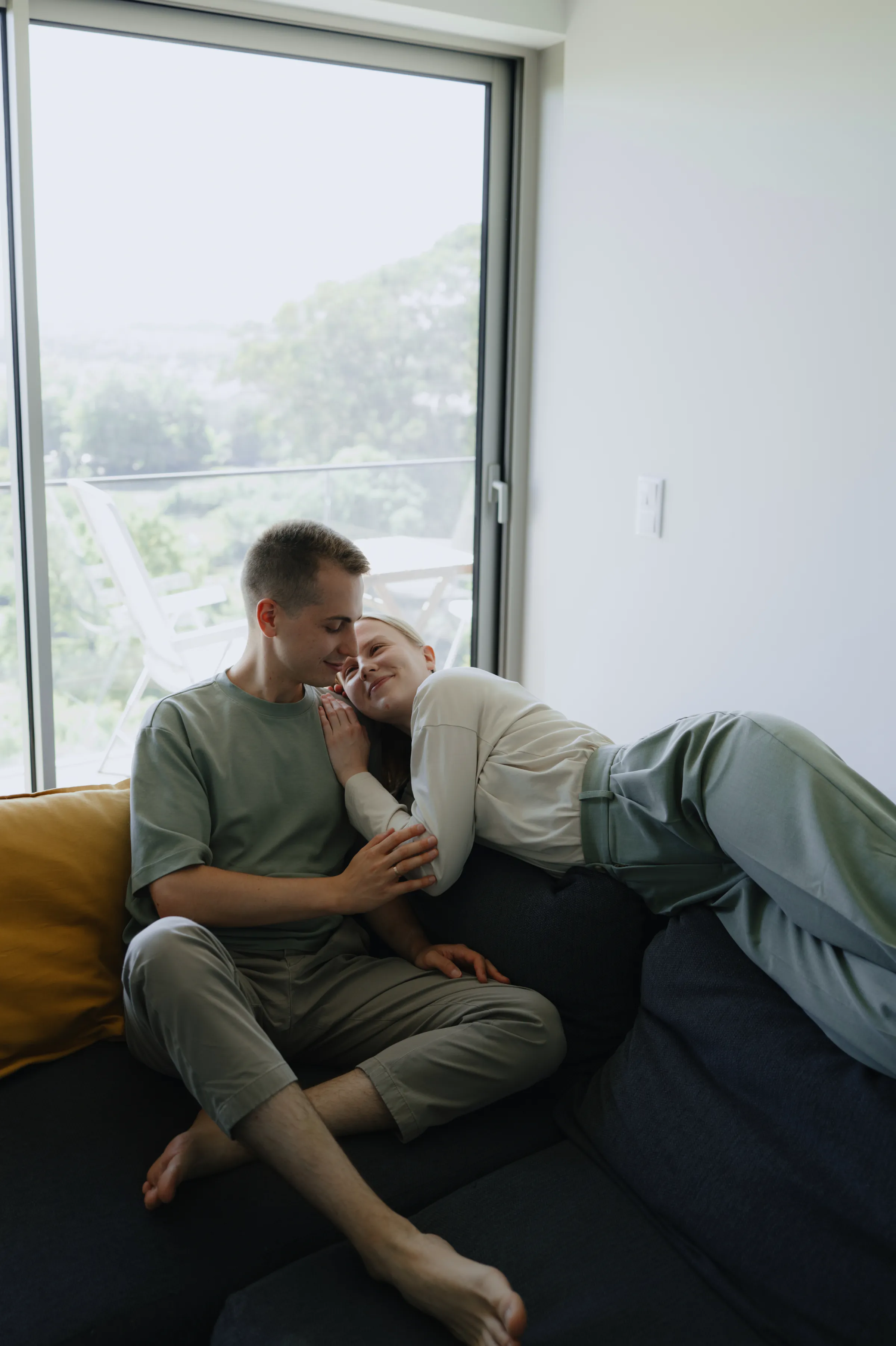 Couple relaxing on a sofa by a large window during a soft light home session.