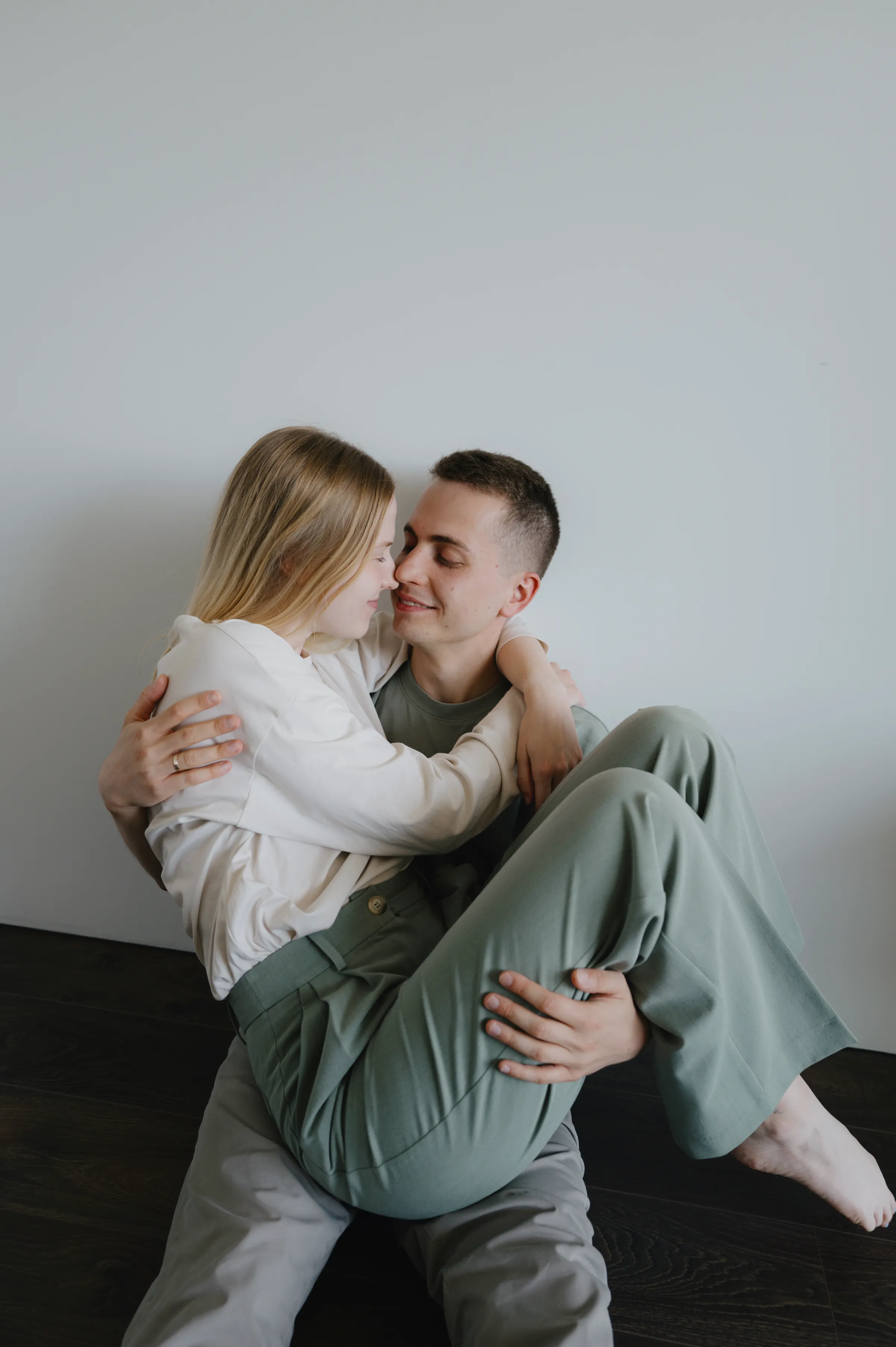 Couple portrait indoors, seated close together against a plain wall in soft light.