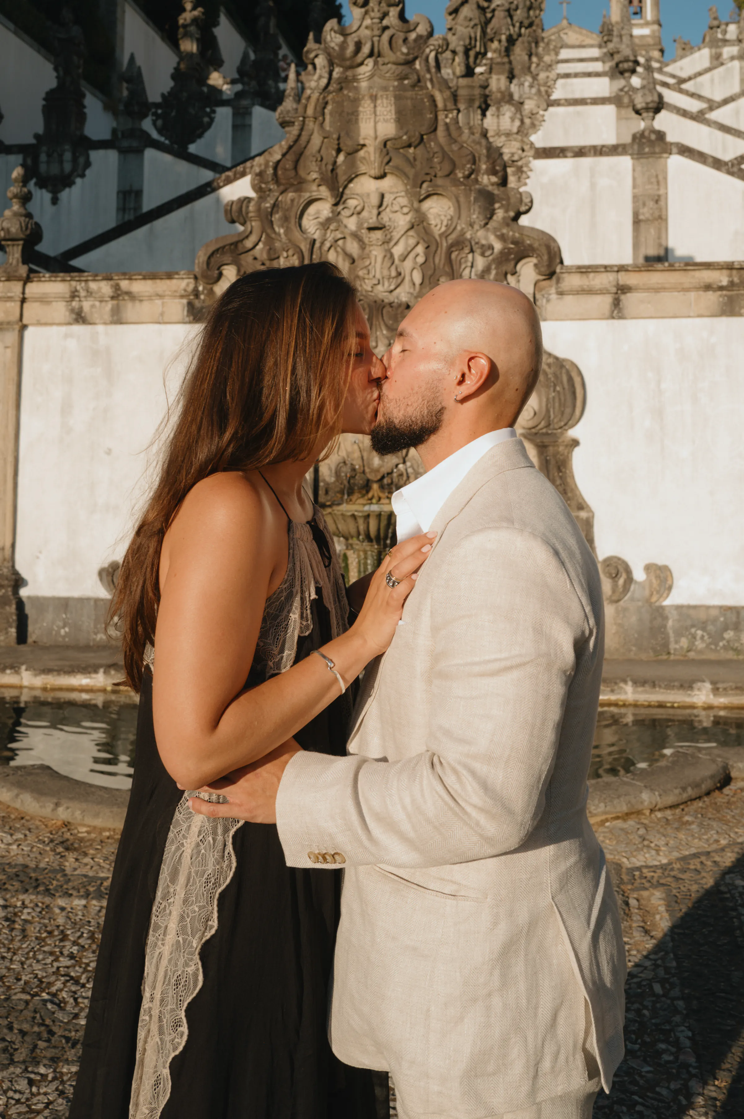 Couple kissing beside an ornate stone fountain during an outdoor city walk in soft light.