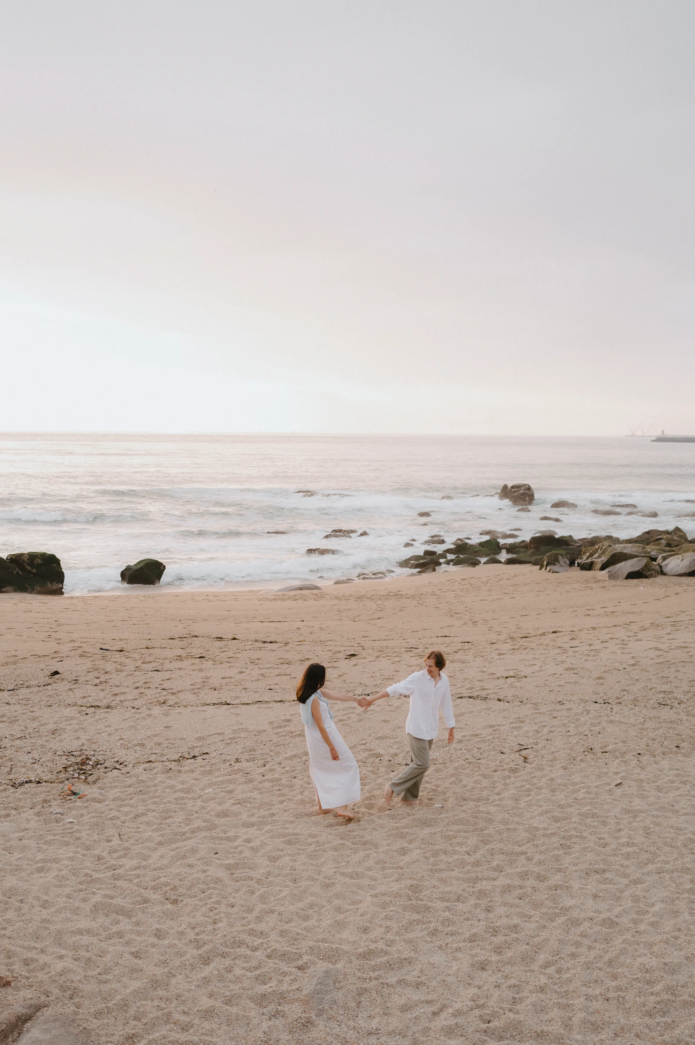 Couple on a sandy seaside outdoor session, standing near calm waves in soft light.