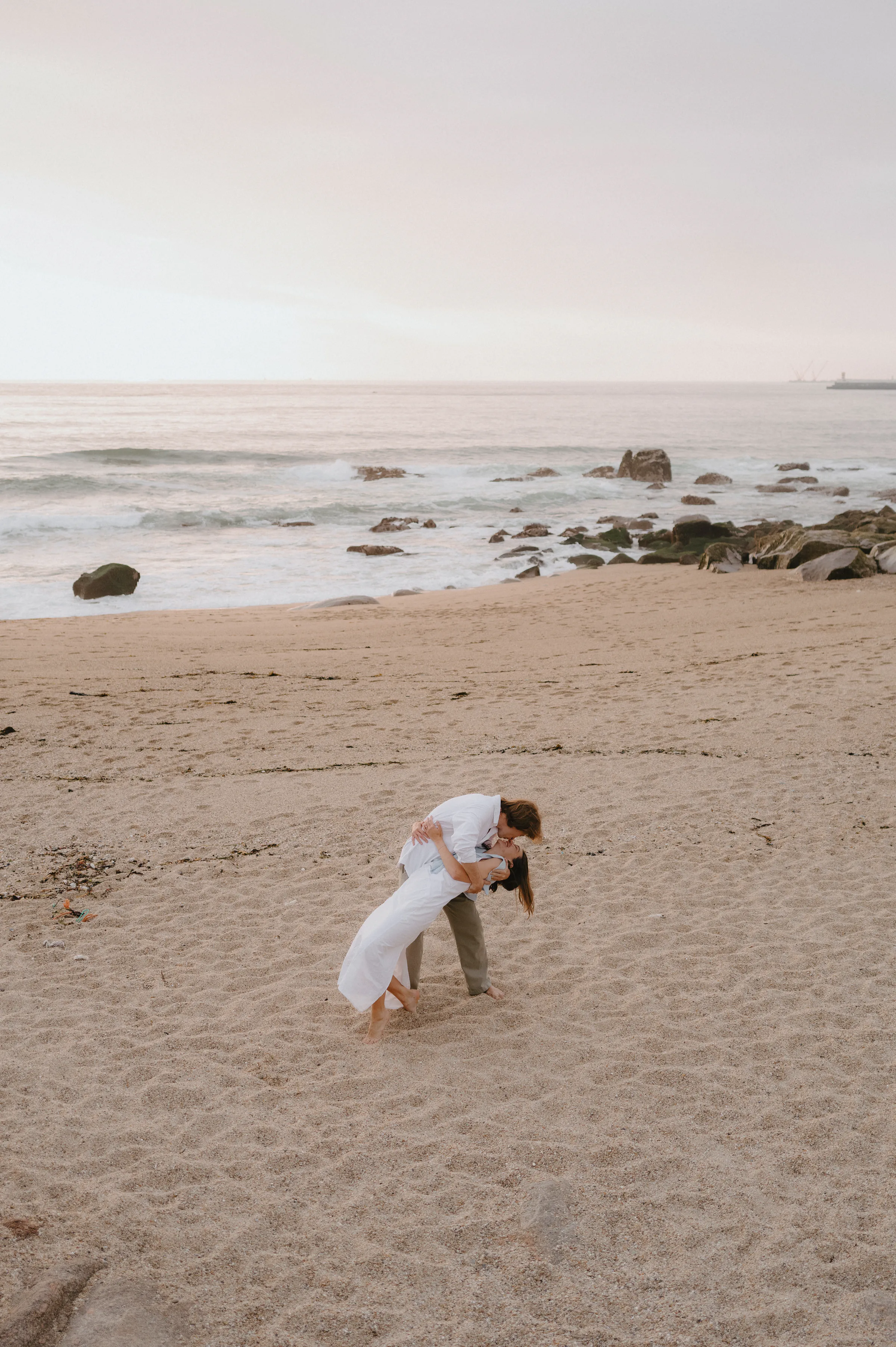 Couple embracing on a sandy seaside during an outdoor session, with rocks, gentle waves, and soft light.