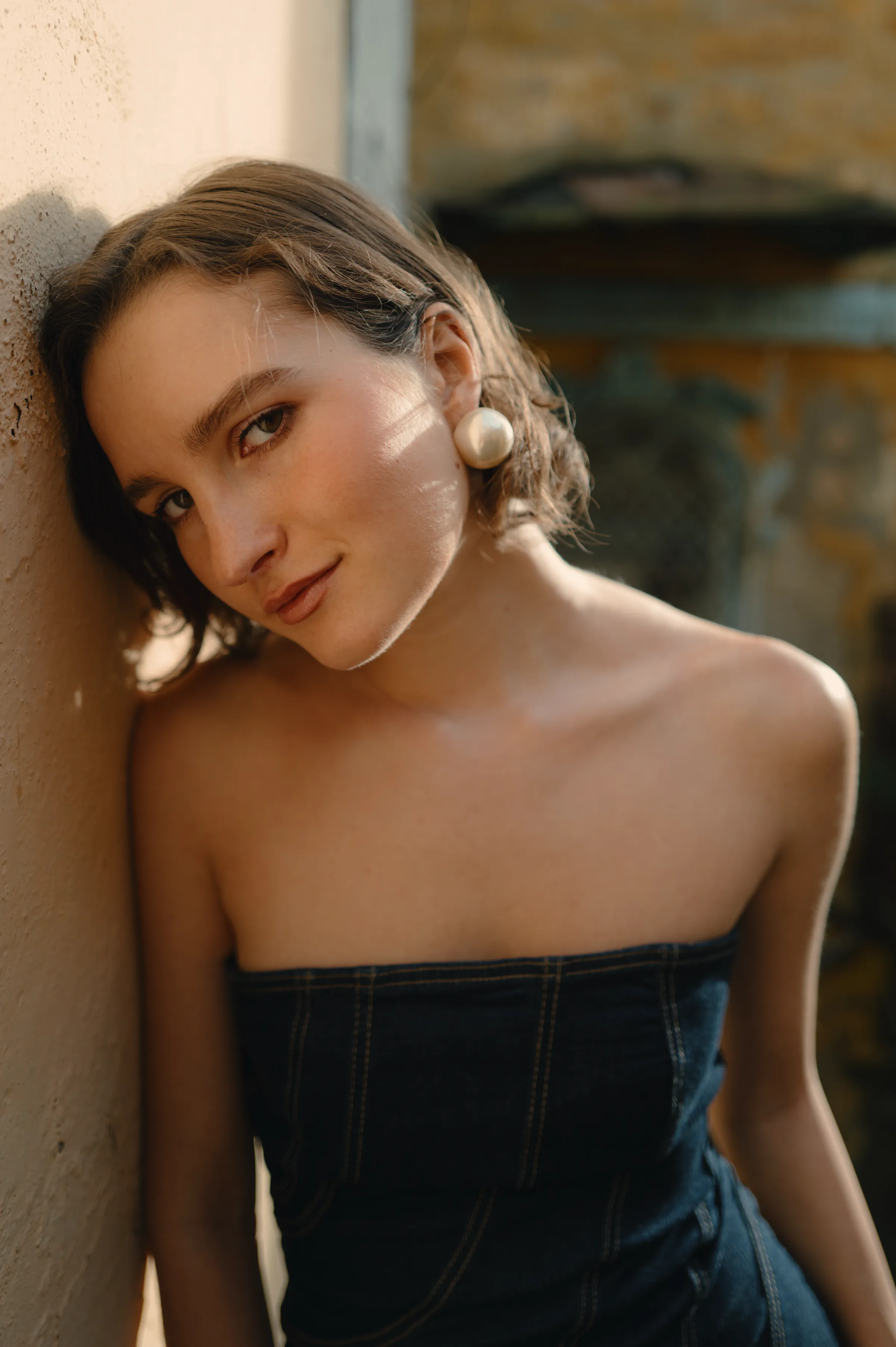Close outdoor portrait of a young adult leaning against a textured wall in soft light.