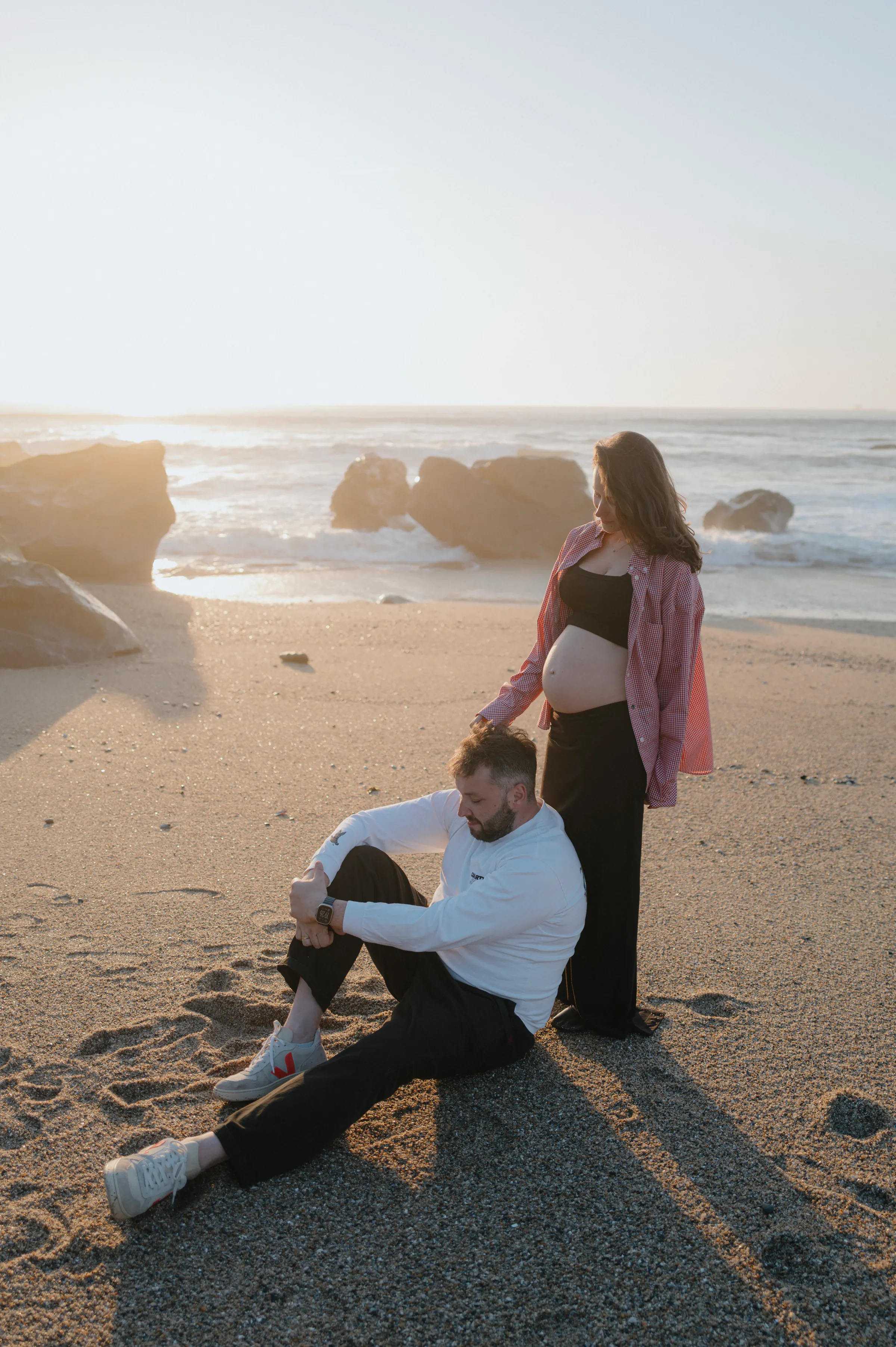 Couple during a seaside outdoor session, one standing with a visible baby bump as waves break behind them.