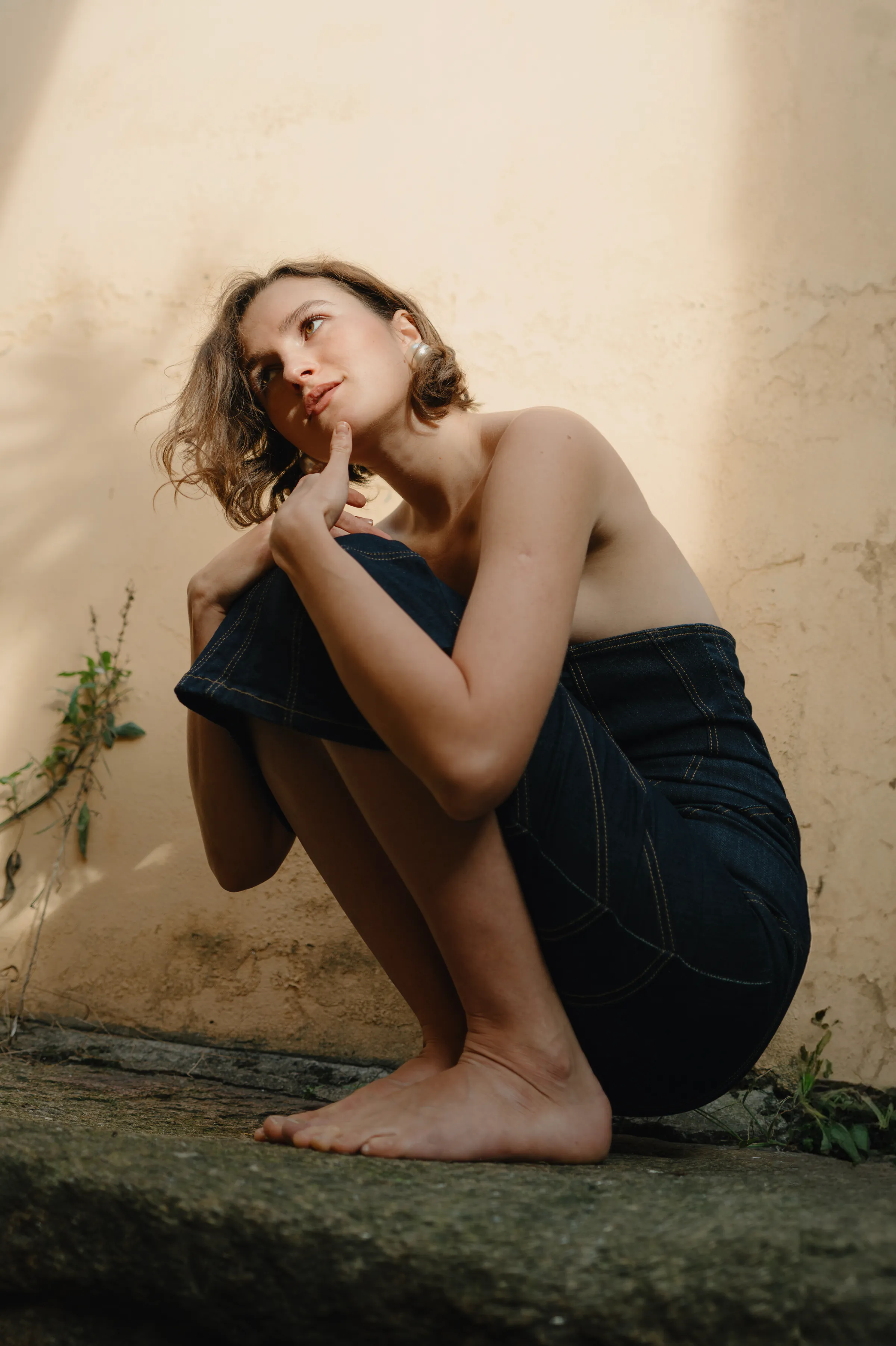 Outdoor portrait of a seated woman in denim, barefoot against a textured wall in soft light