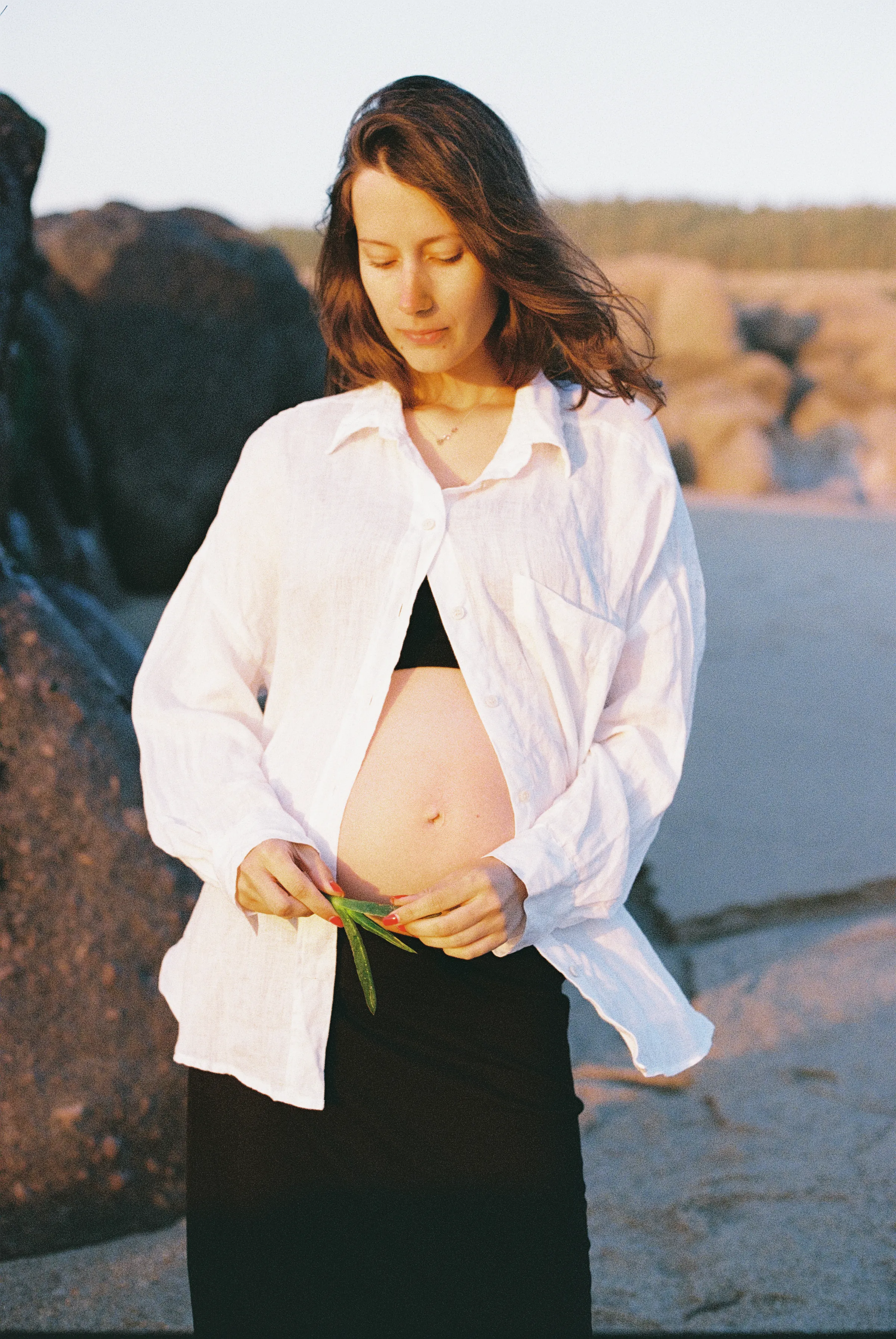 Outdoor portrait by rocky seaside, with a person in a white shirt holding a small flower in soft light.