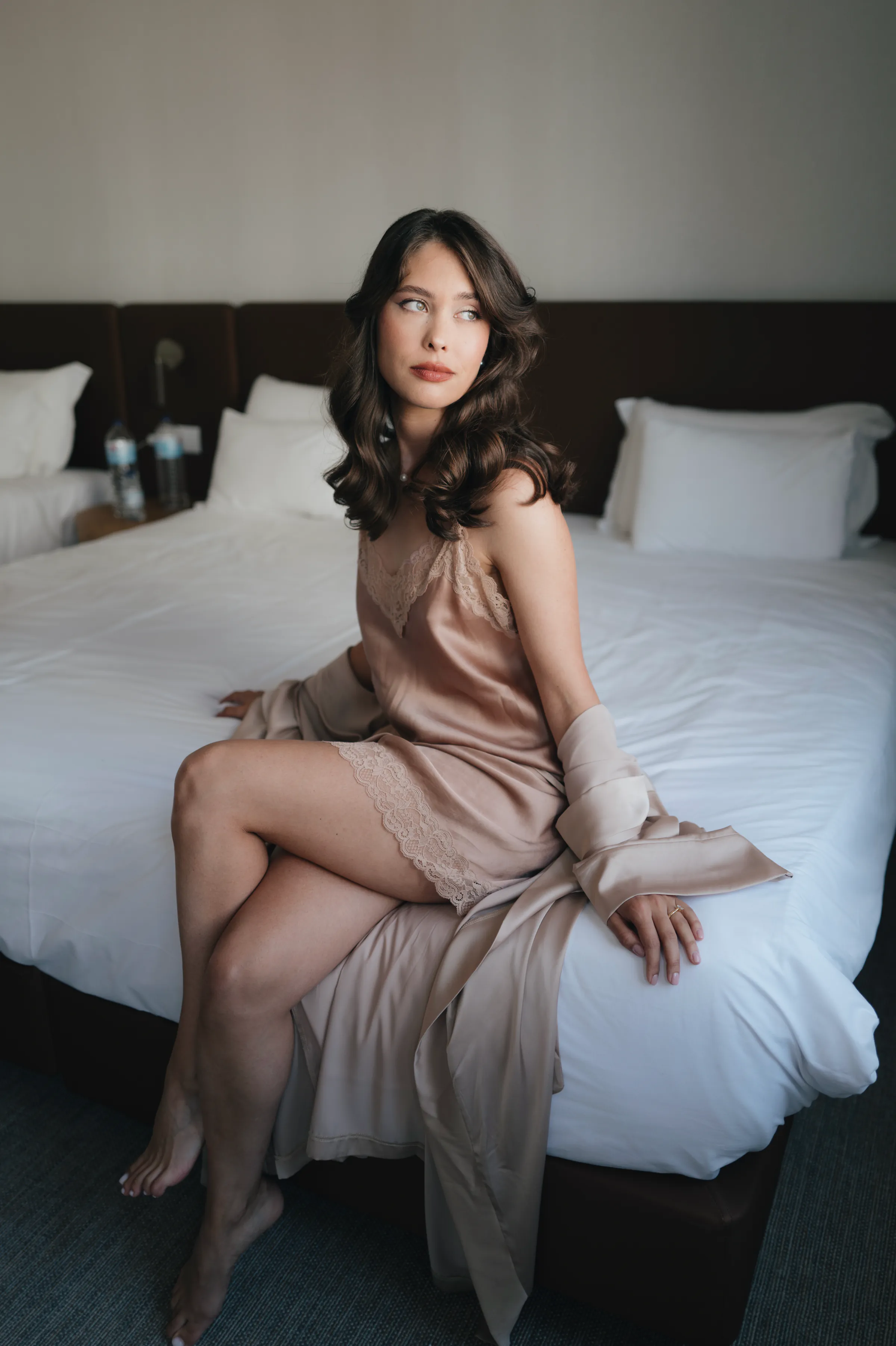 Portrait of a woman seated on a bed in soft indoor light, wearing a satin dress and gloves.