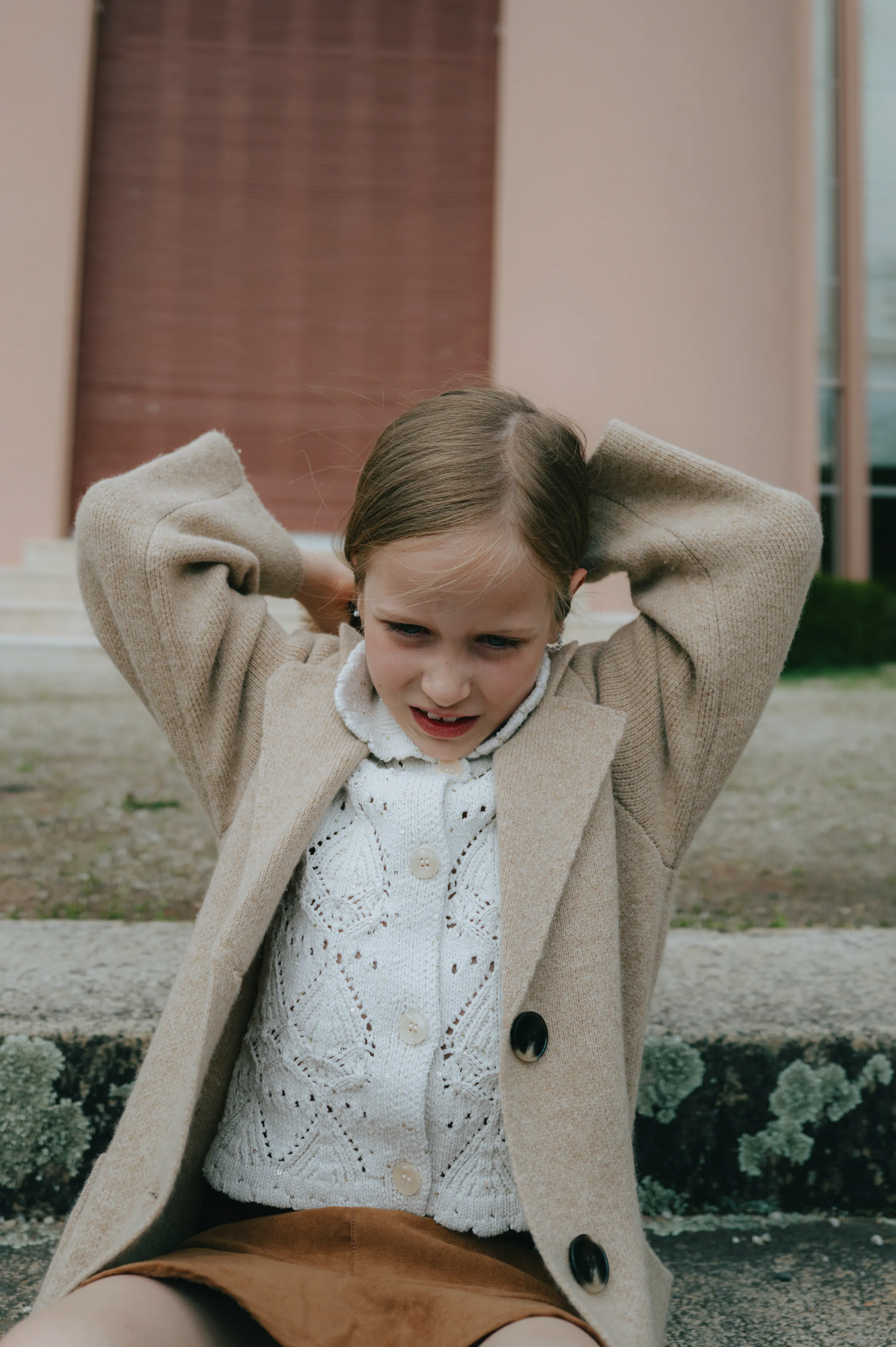 Child portrait on outdoor steps, wearing a beige cardigan with hands tucked behind their head.