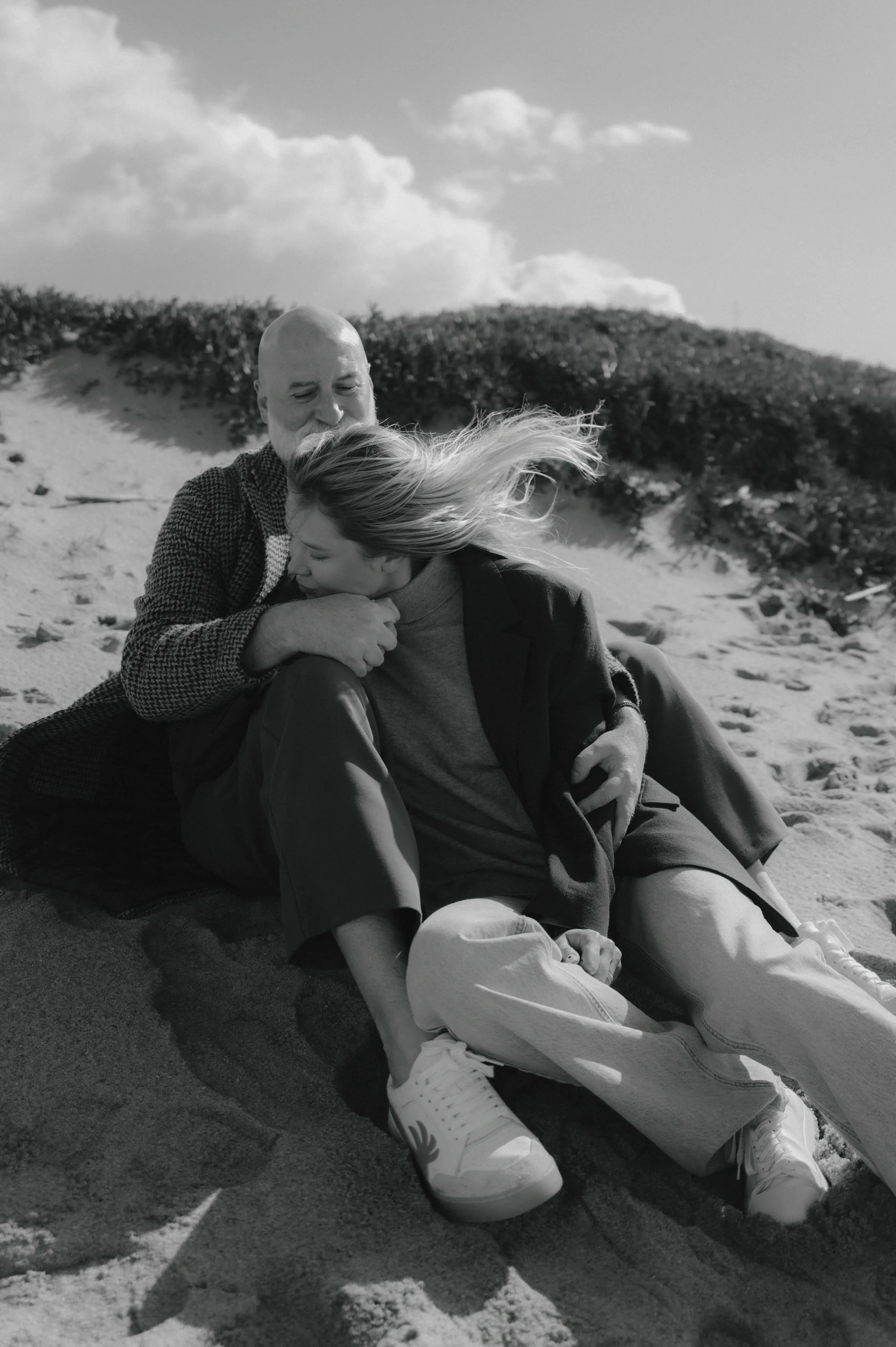 Black-and-white outdoor session portrait of a couple sitting close together on sandy dunes in soft light.
