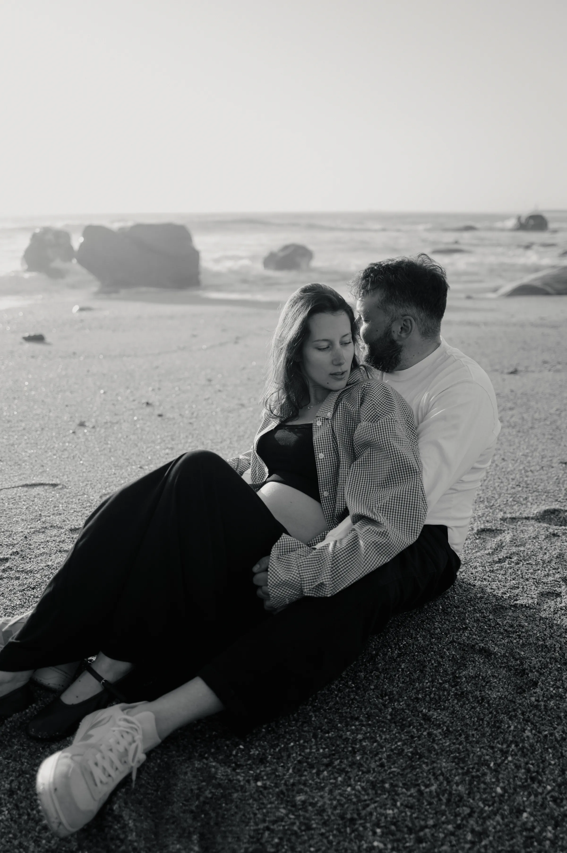 Black-and-white seaside portrait of a couple seated close together on sandy beach rocks in soft light.
