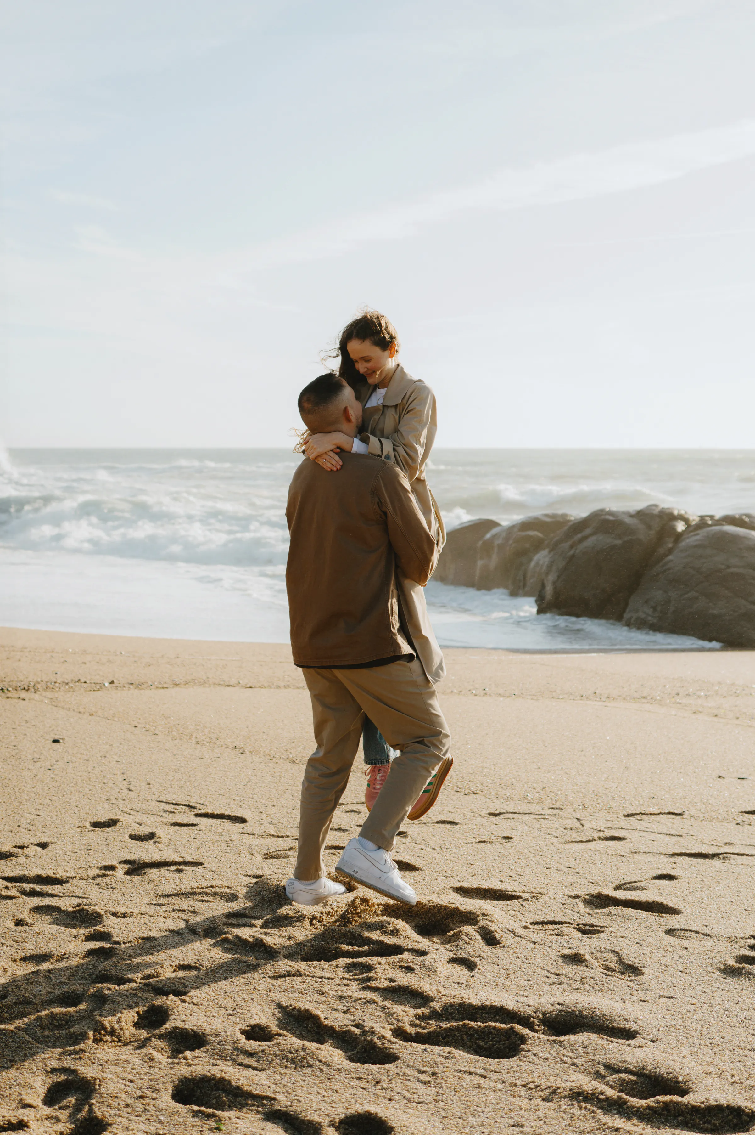 Couple during a seaside session, one person carrying the other on a sandy beach near rocks and surf.