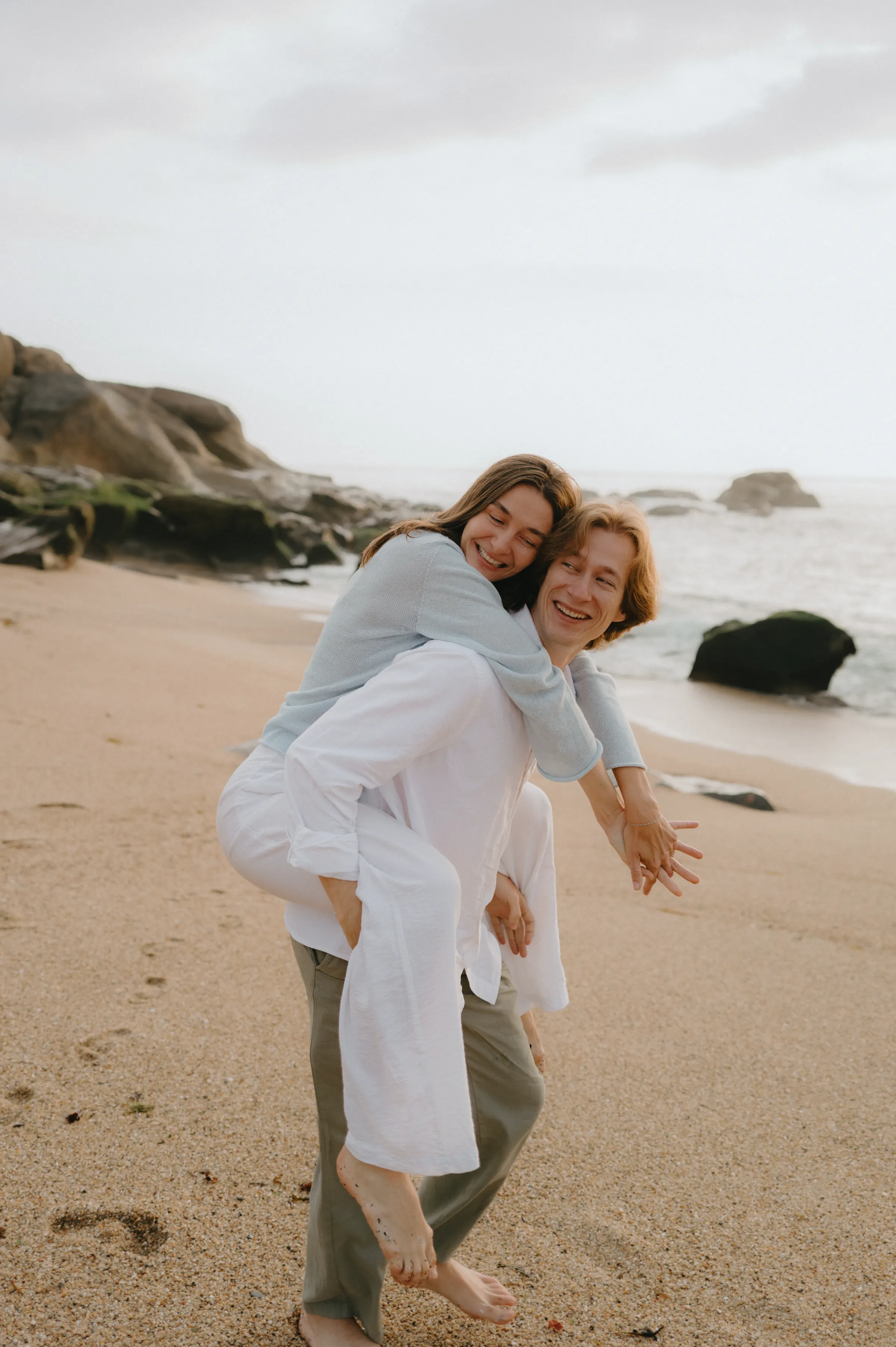 Couple during a seaside session, one giving the other a piggyback ride on a sandy beach.