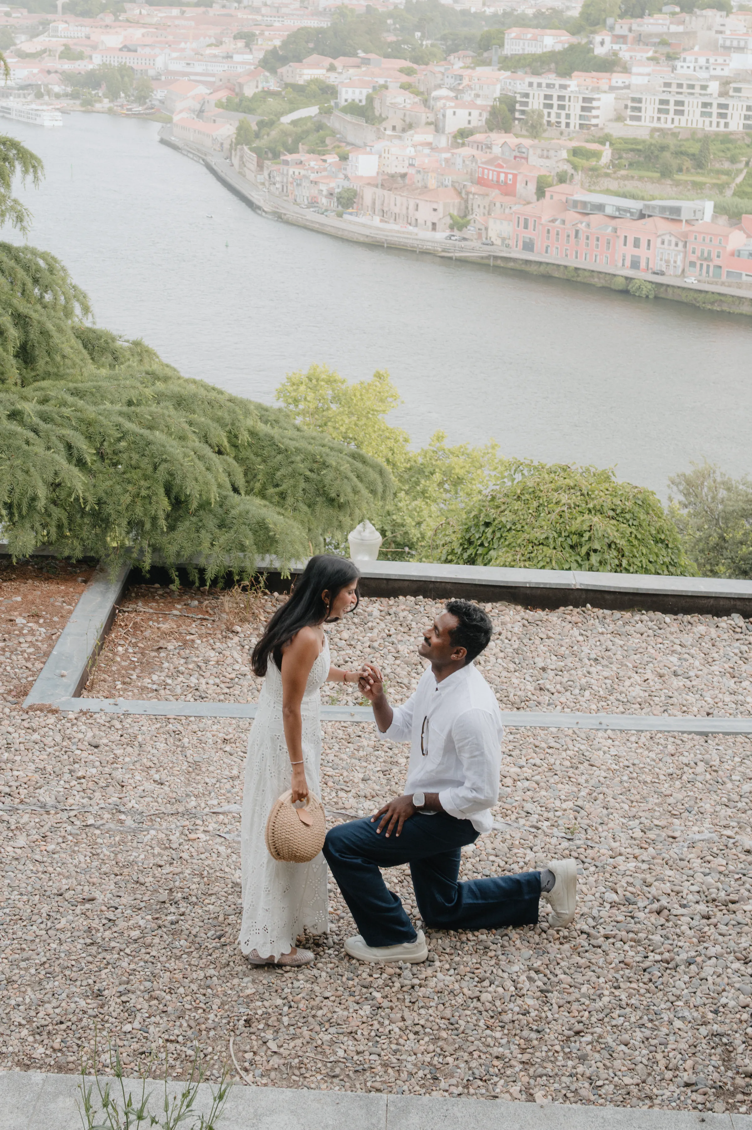 Couple during an outdoor session on a gravel overlook, one kneeling beside a river and city backdrop.