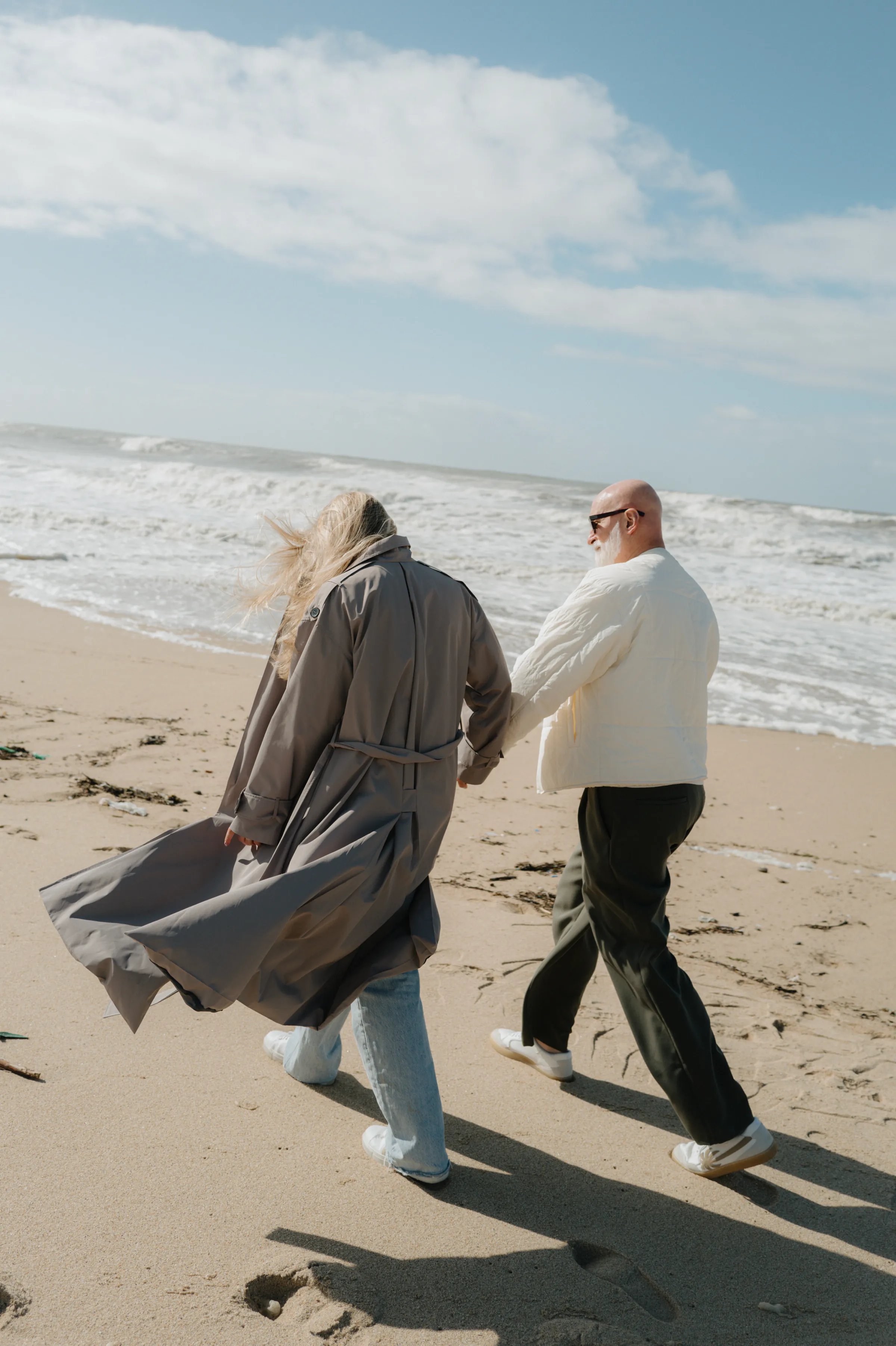Couple walking hand in hand during a seaside outdoor session, with windblown coats and waves behind them.