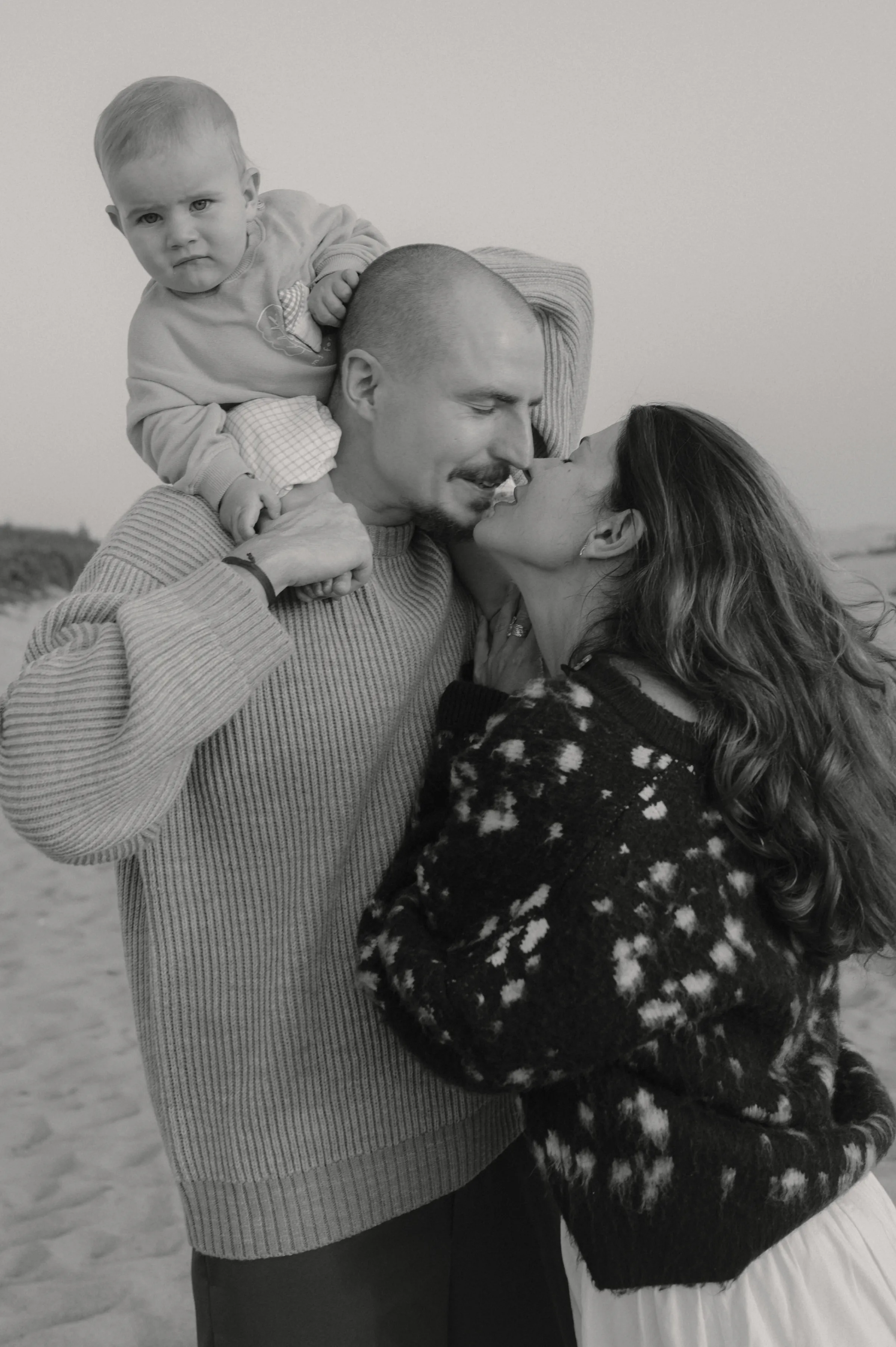 Black-and-white seaside family portrait with a baby on an adult's shoulders as the couple shares a close kiss.