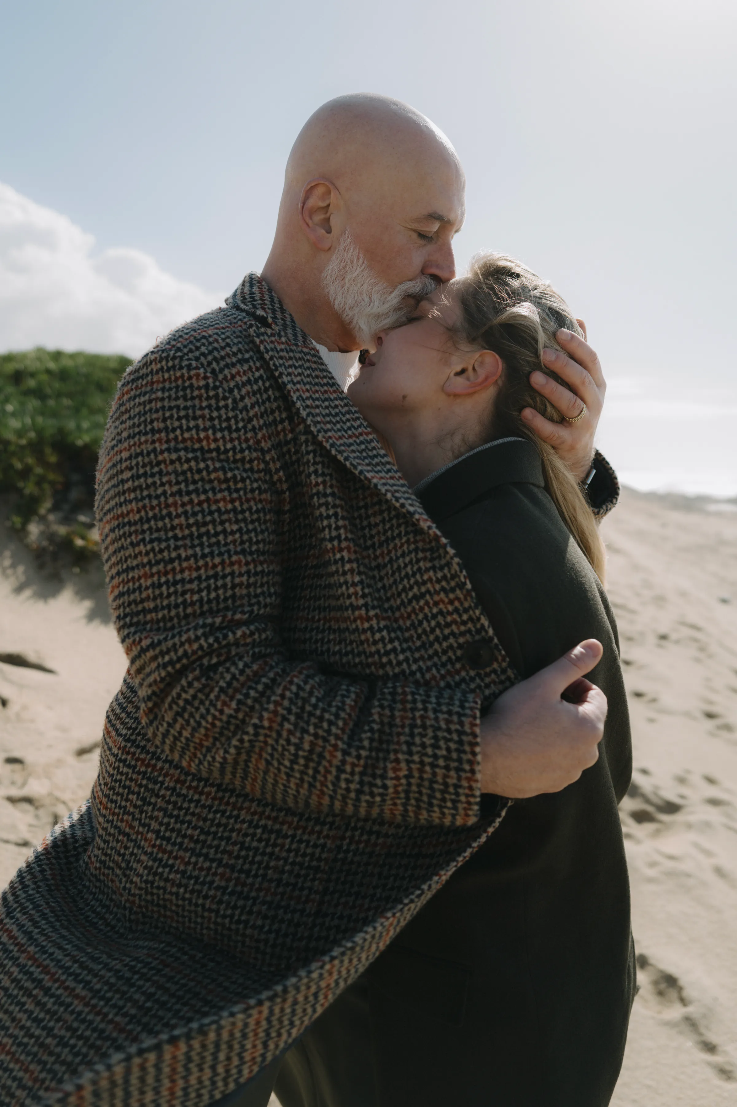 Couple embracing during a seaside outdoor session, standing close on a sandy beach in soft light.
