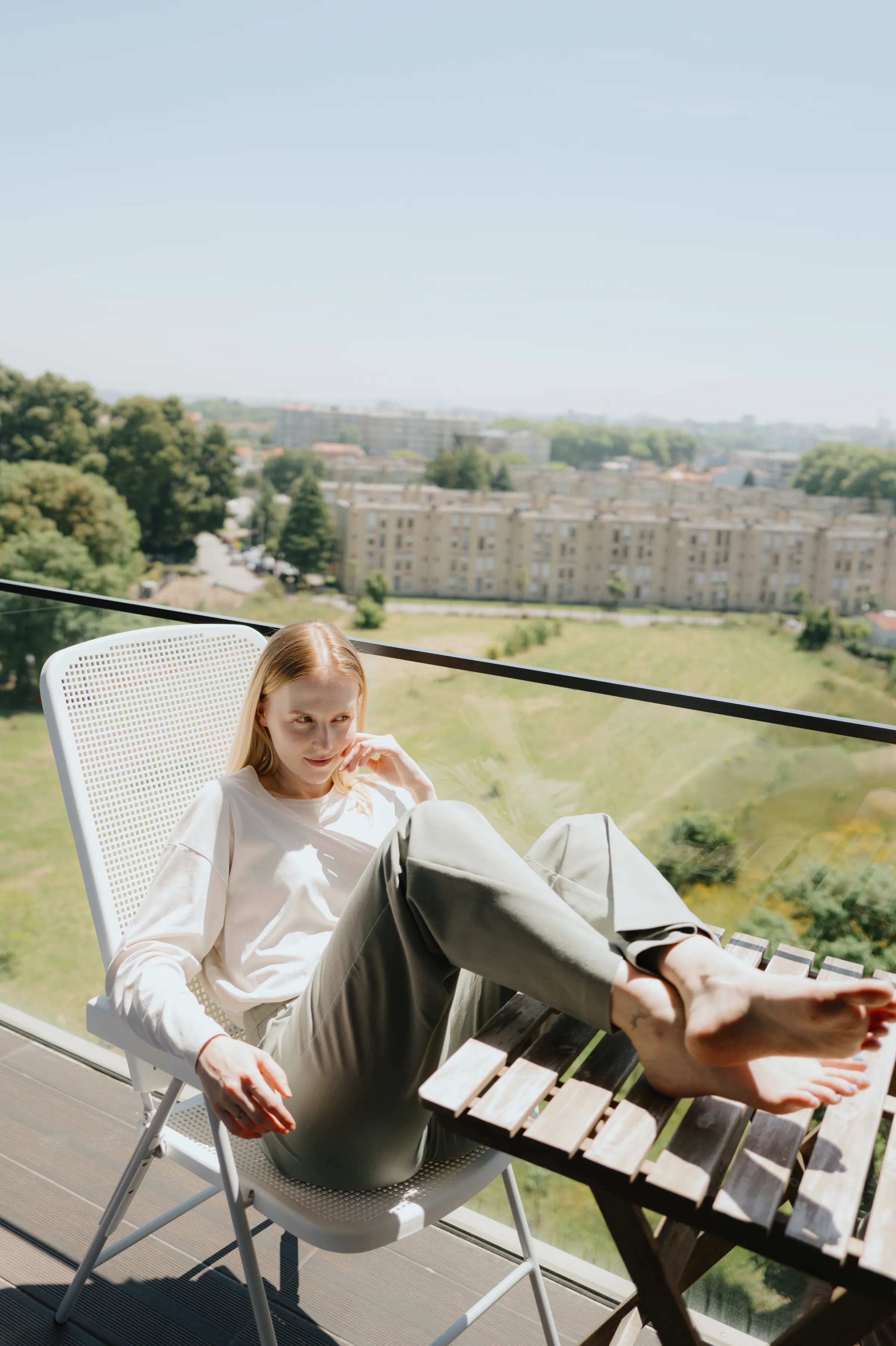 Outdoor portrait on a balcony, with a seated person overlooking green space and city buildings in soft daylight.