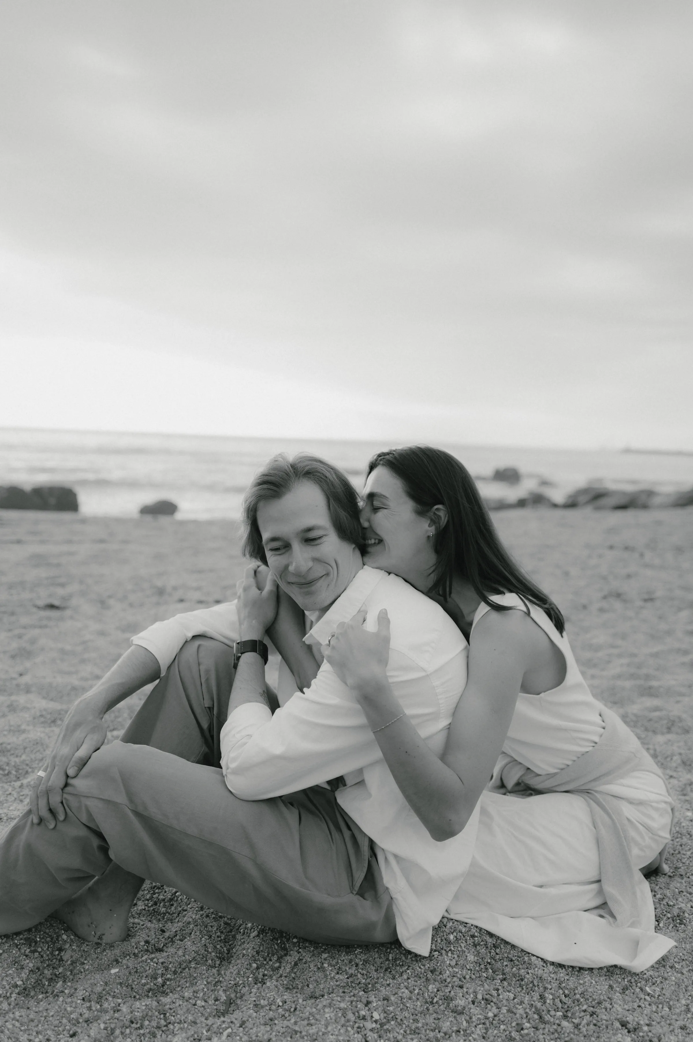 Black-and-white seaside portrait of a couple seated close together on sand under an overcast sky.