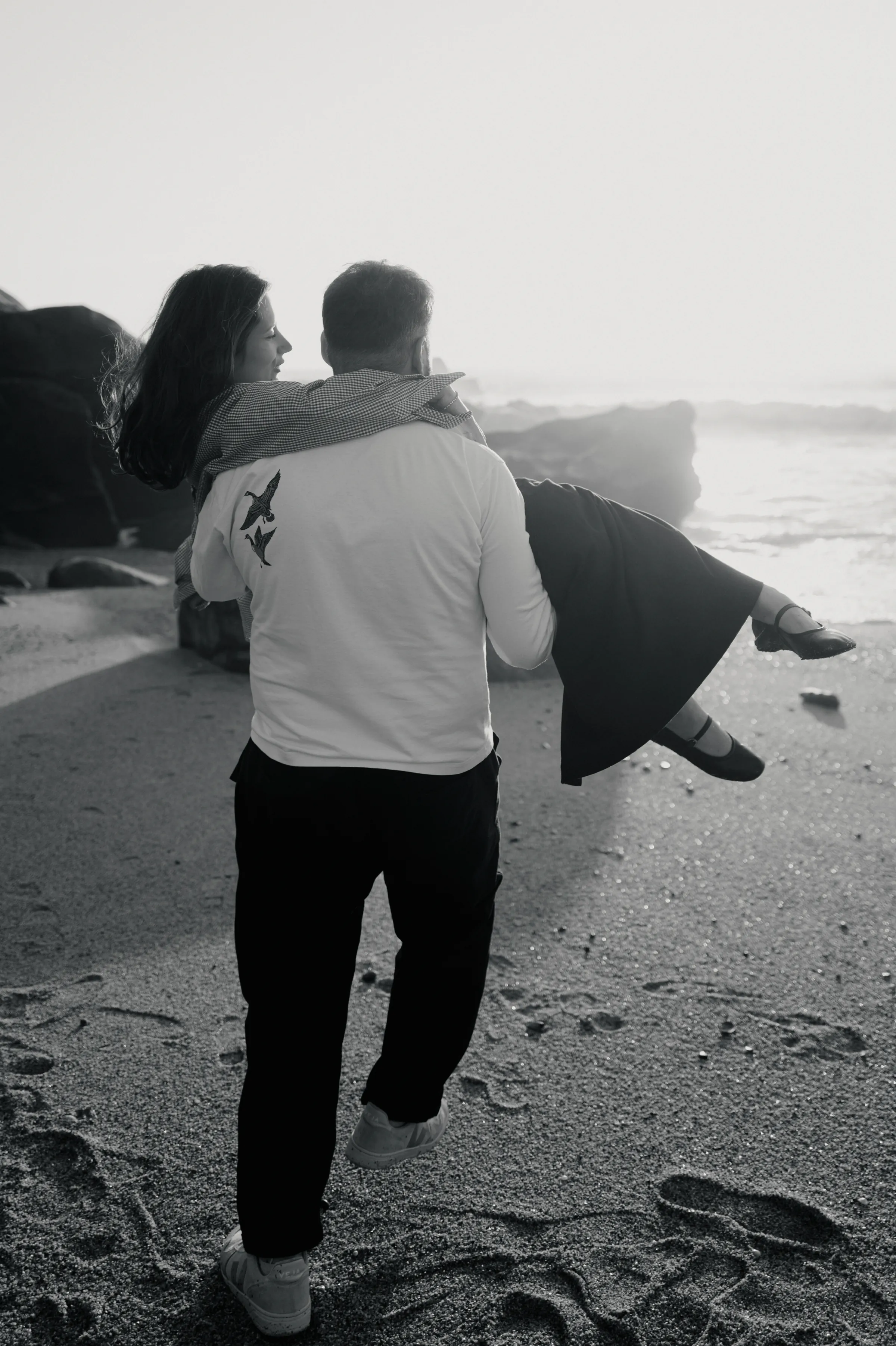 Black-and-white seaside portrait of a couple, one carrying the other along a sandy beach near the surf.