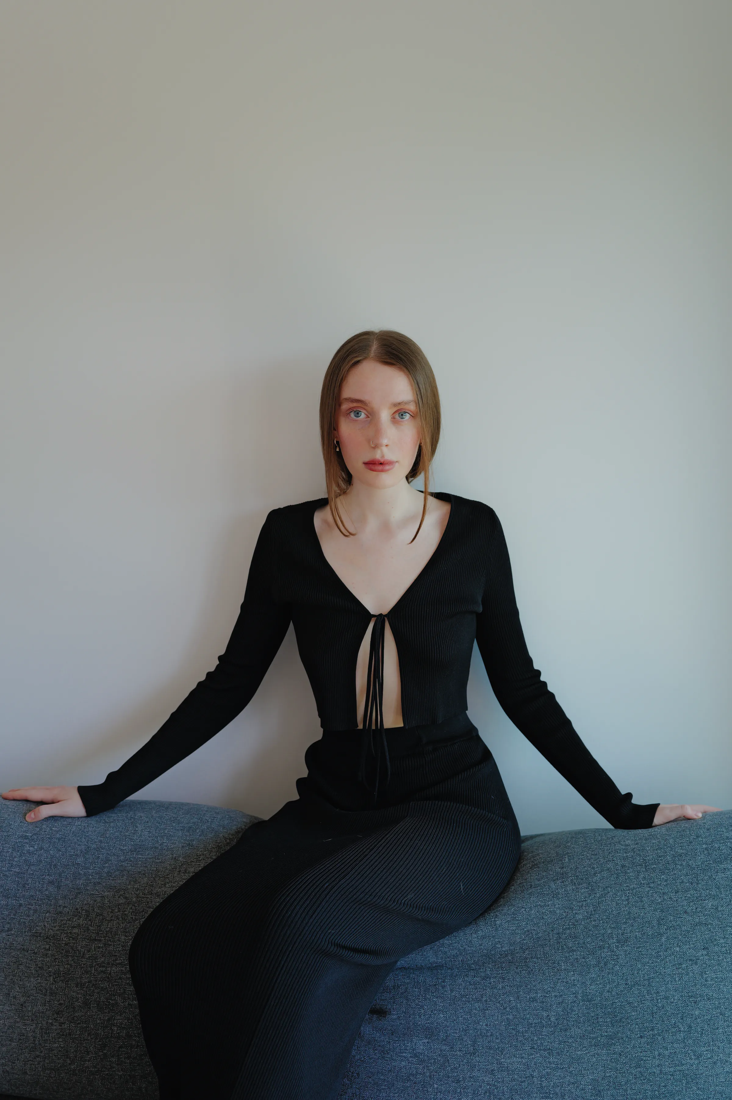 Indoor portrait of a seated woman on a gray sofa against a plain wall, in soft light.