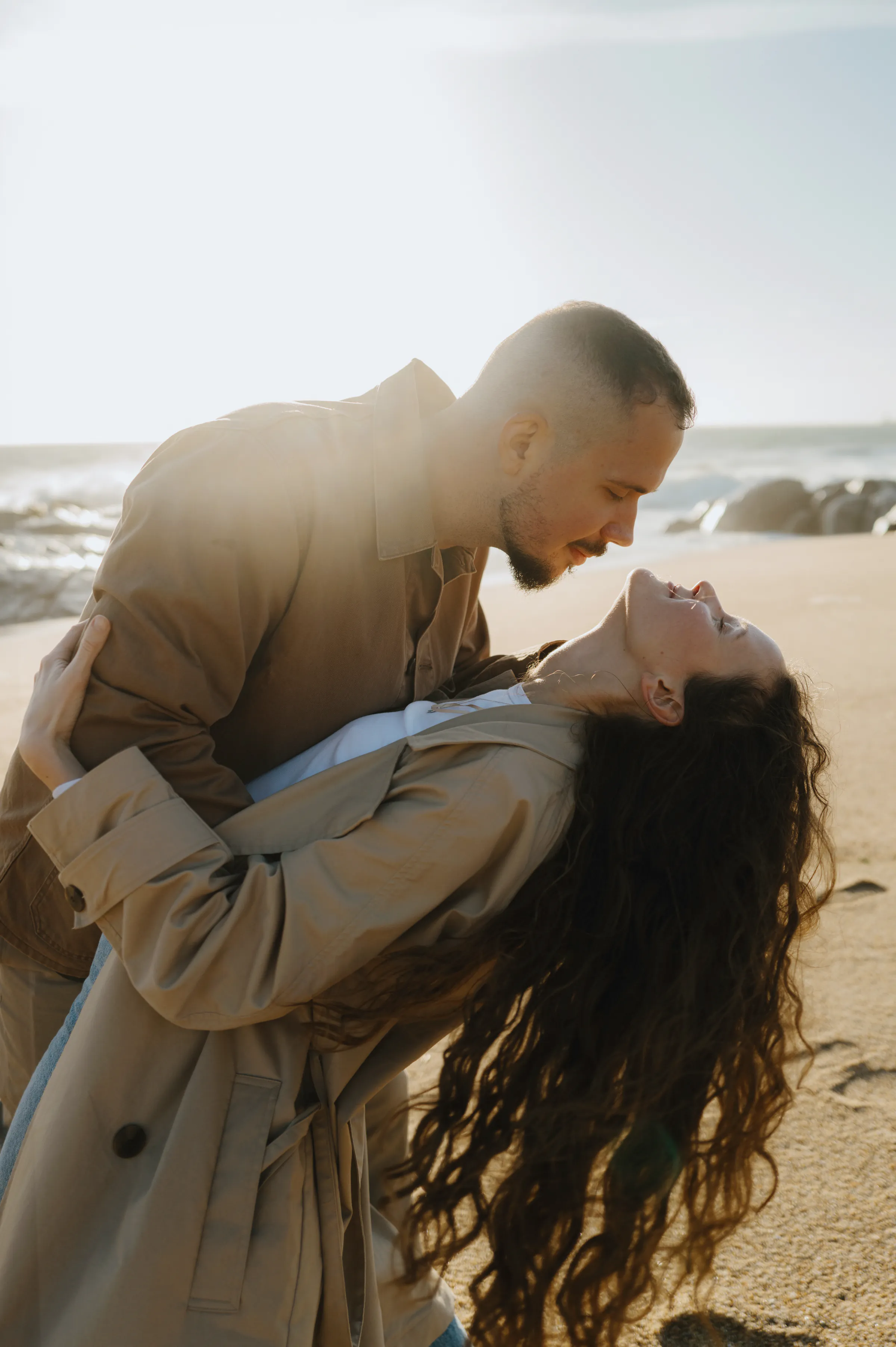 Couple in beige coats leaning together during a seaside portrait session in soft backlight