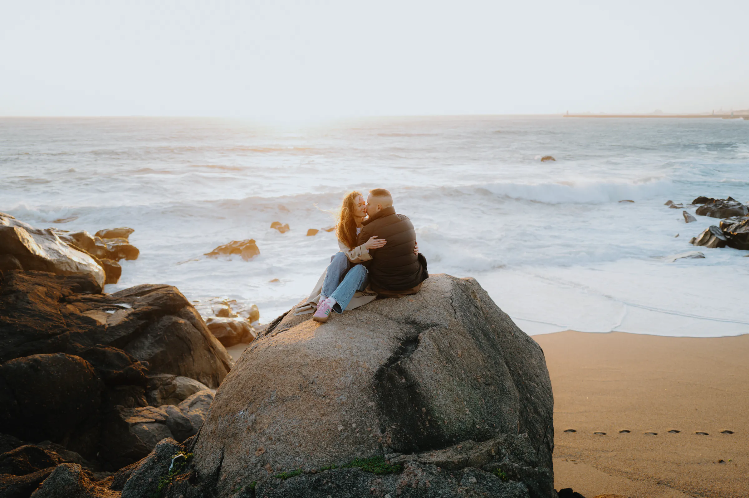 Couple seated on a large seaside rock, embracing during a soft light outdoor session by the waves.