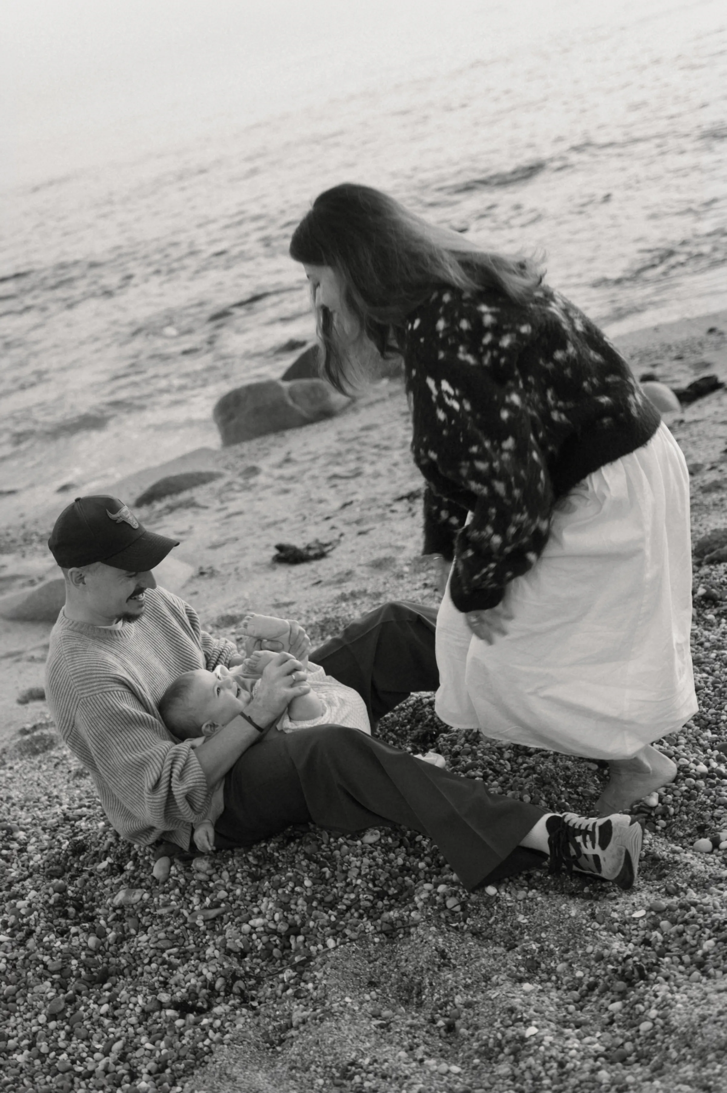 Black-and-white seaside family portrait with one adult holding a baby as another leans close on a pebble beach.