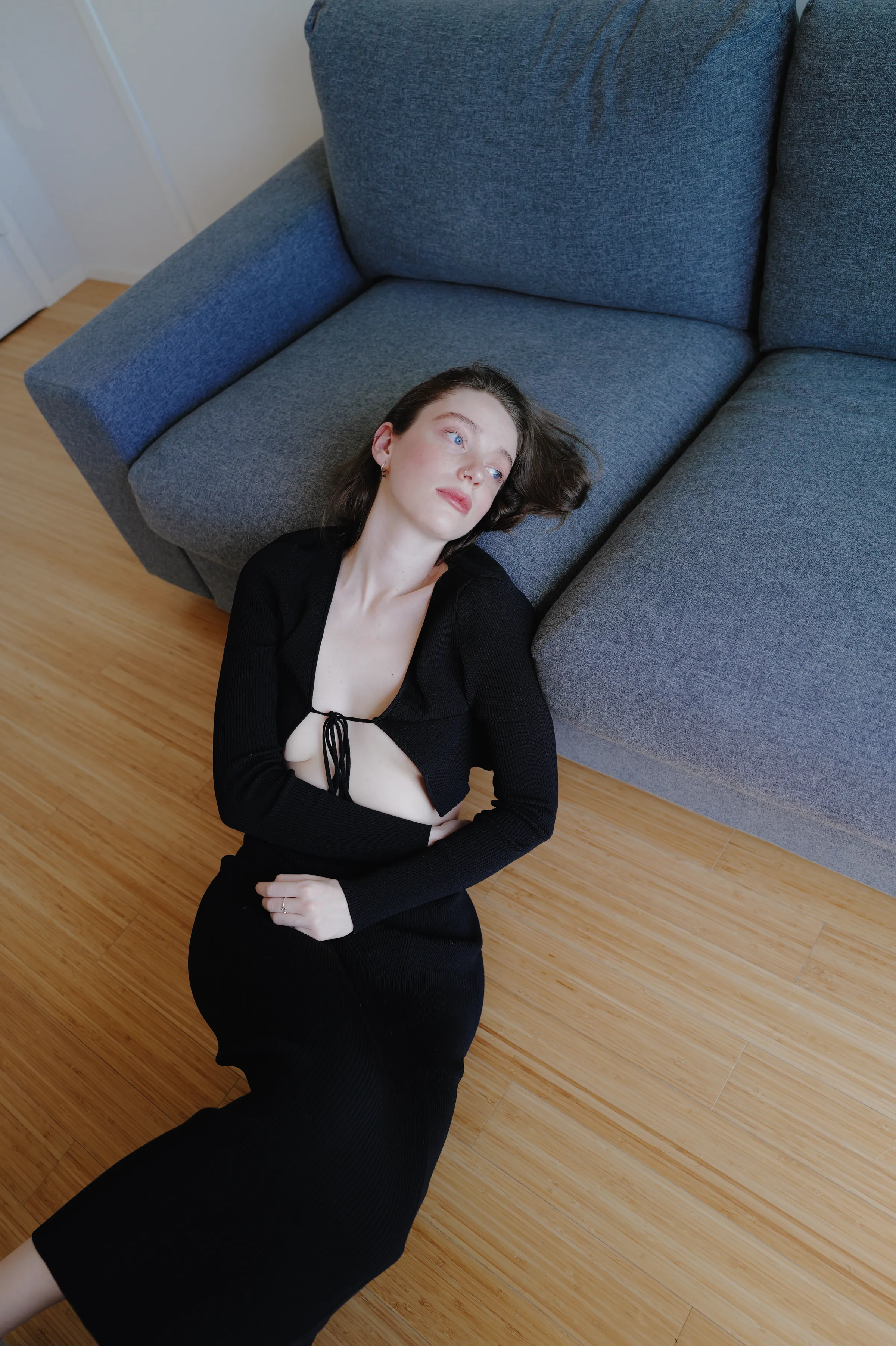Overhead indoor portrait of a woman in black reclining on a wood floor beside a gray sofa.