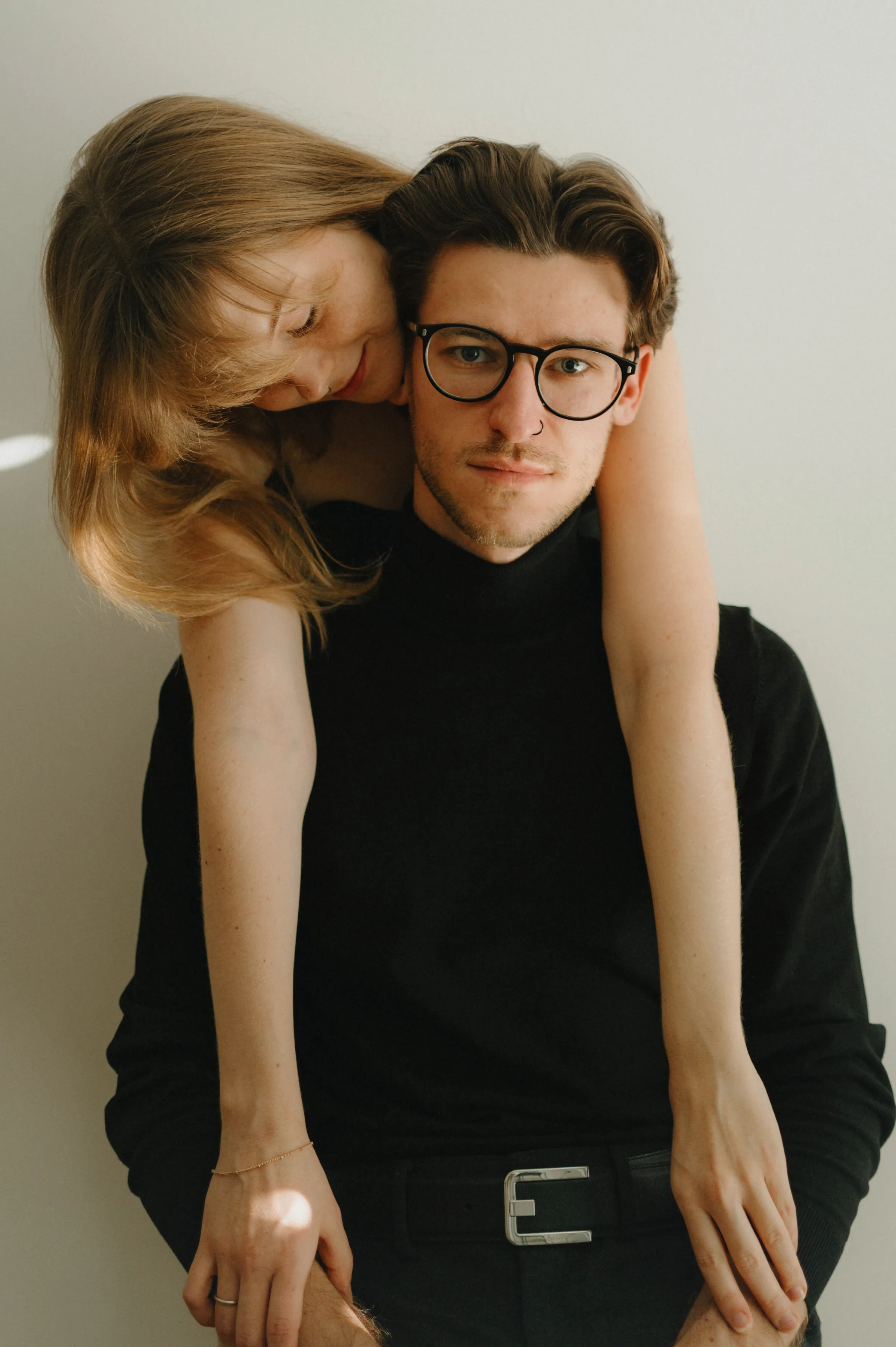 Close portrait of a couple indoors, one leaning over the other's shoulders in soft light.