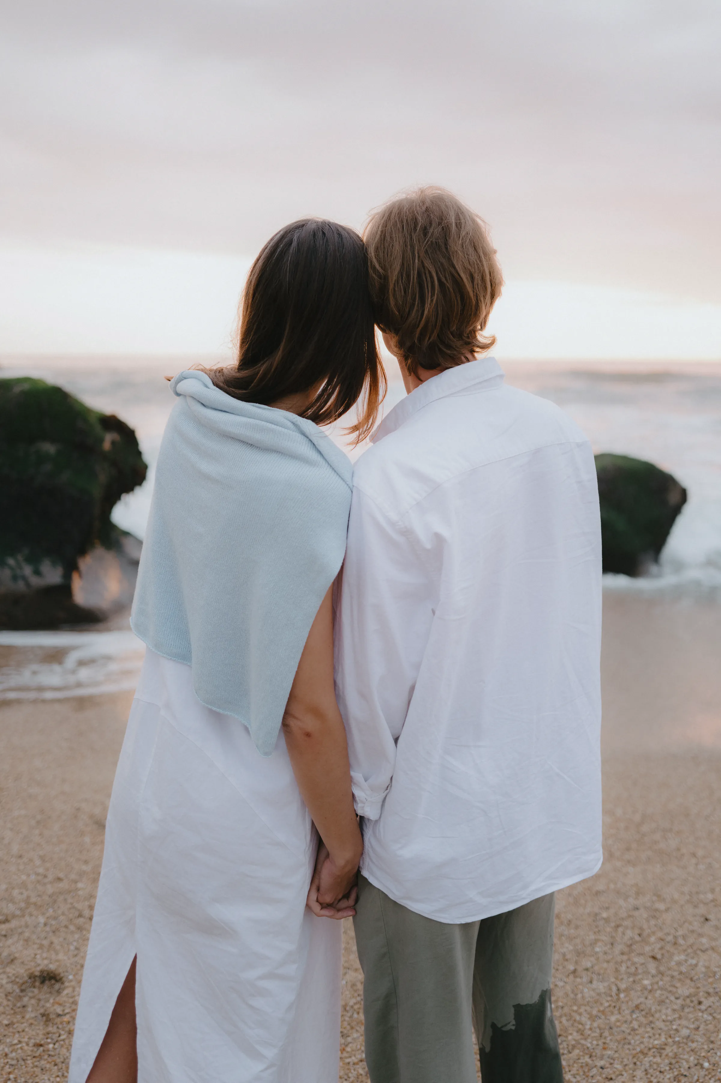 Couple holding hands during a soft light seaside portrait, seen from behind on a sandy beach.