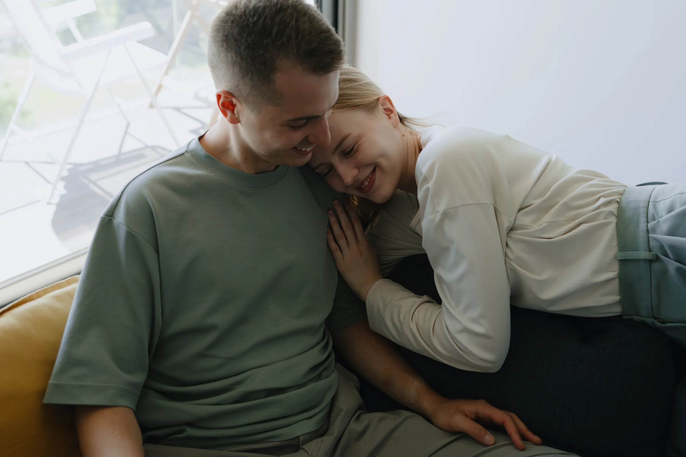 Close indoor couple portrait on a couch by a bright window, captured in soft light.
