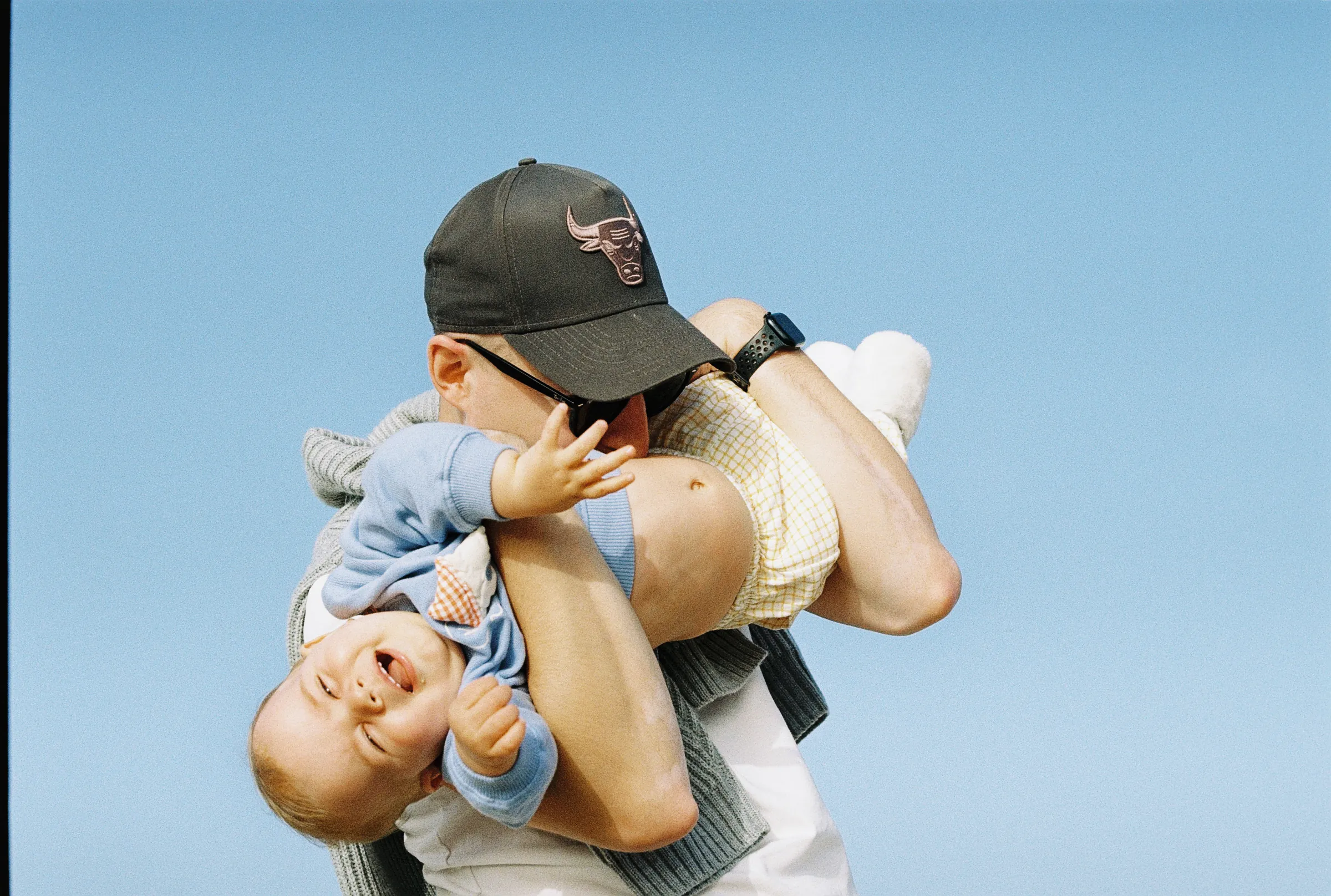 Family outdoor session with an adult in a cap holding a baby against a clear blue sky.