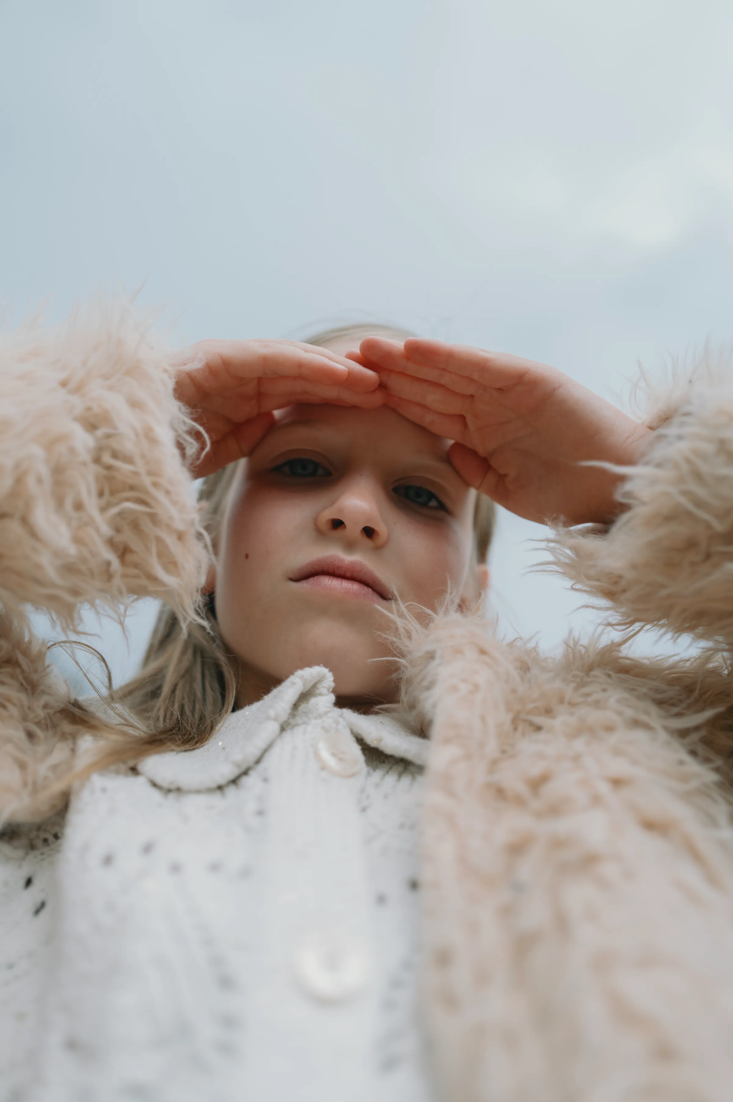 Low-angle close portrait of a person in a textured coat, hand shading their face under soft sky.