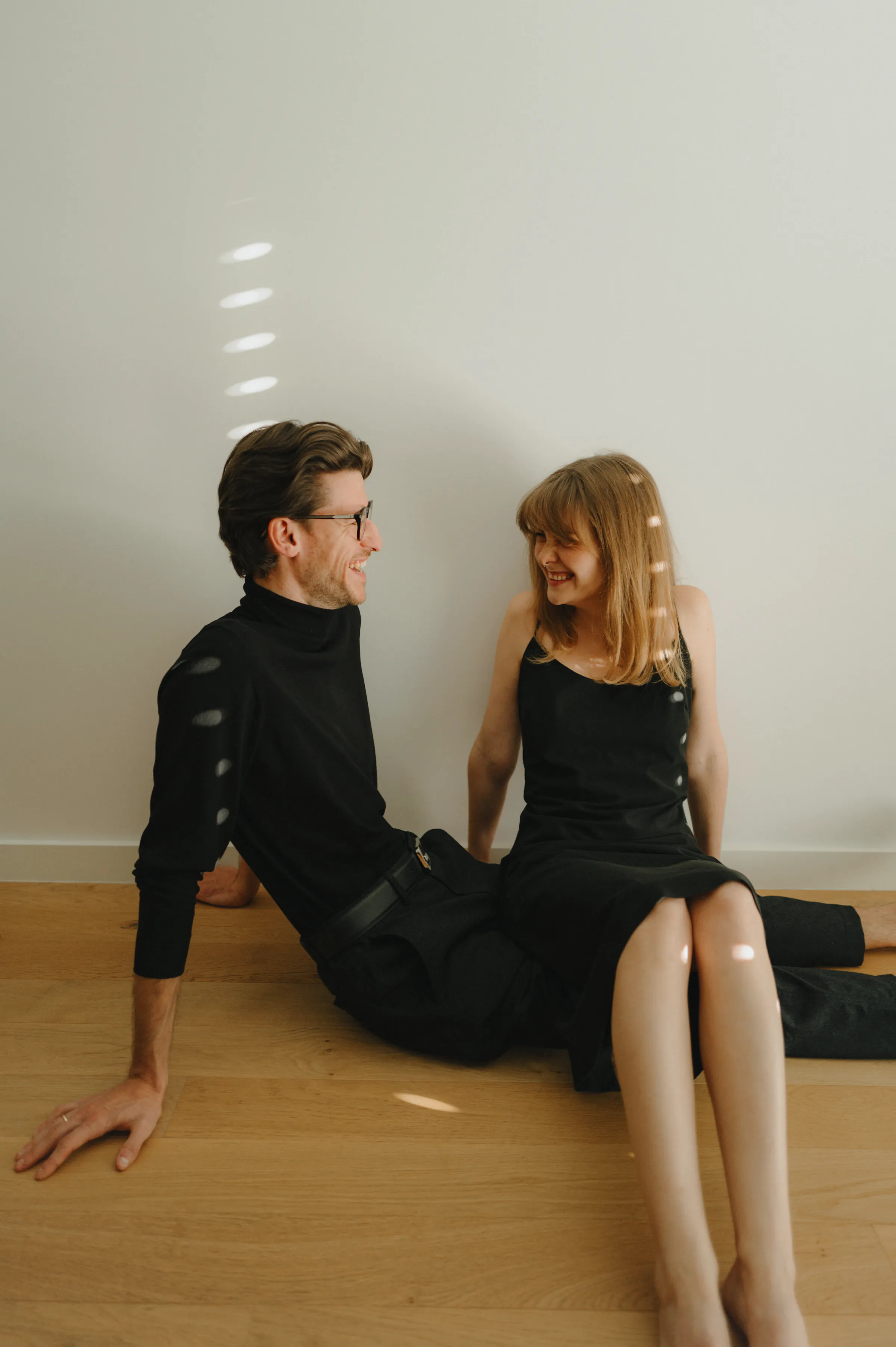 Indoor couple portrait seated on a wooden floor, wearing black outfits in soft striped light.