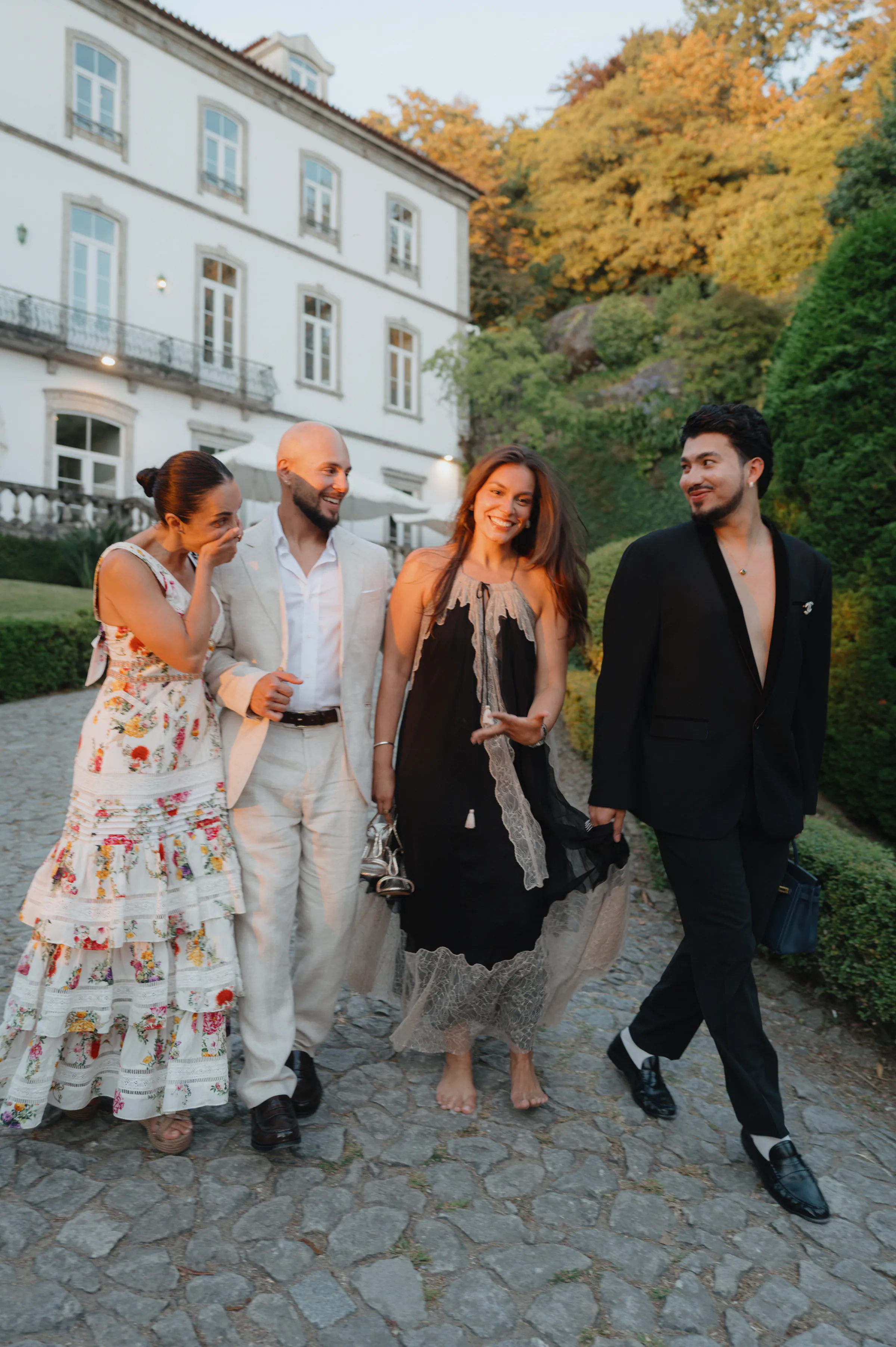 Four adults in formal outfits walk on a cobblestone path during an outdoor session by a white building.
